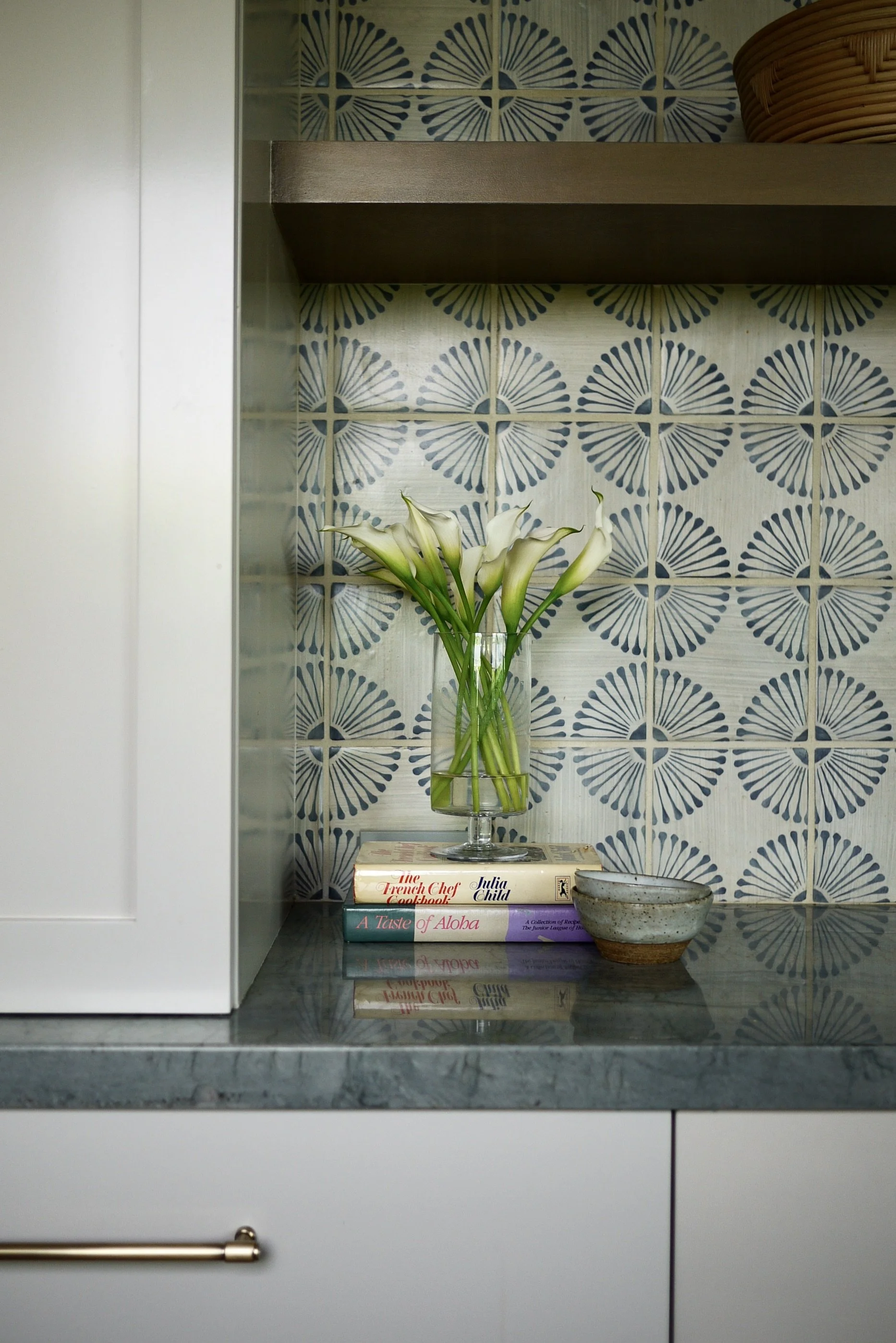 A kitchen counter with a vase of white calla lilies on top of a stack of cookbooks against a blue and white patterned backsplash.