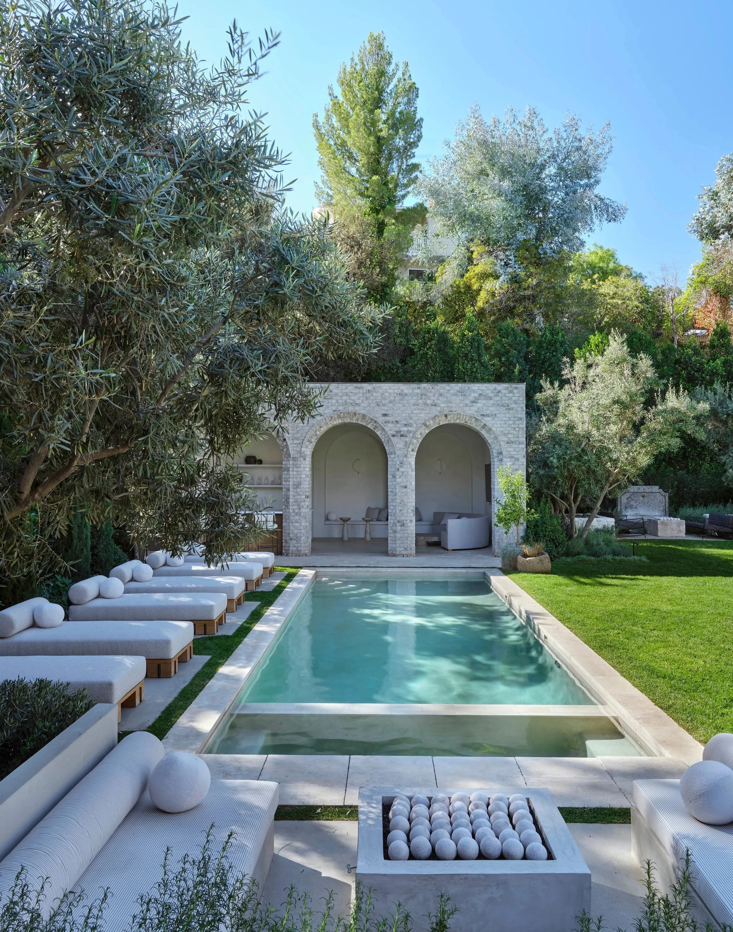 Outdoor view of a luxurious backyard with a rectangular swimming pool surrounded by cushioned benches. A building with stone arches and a sitting area is visible in the background. The area is landscaped with green trees and grass.