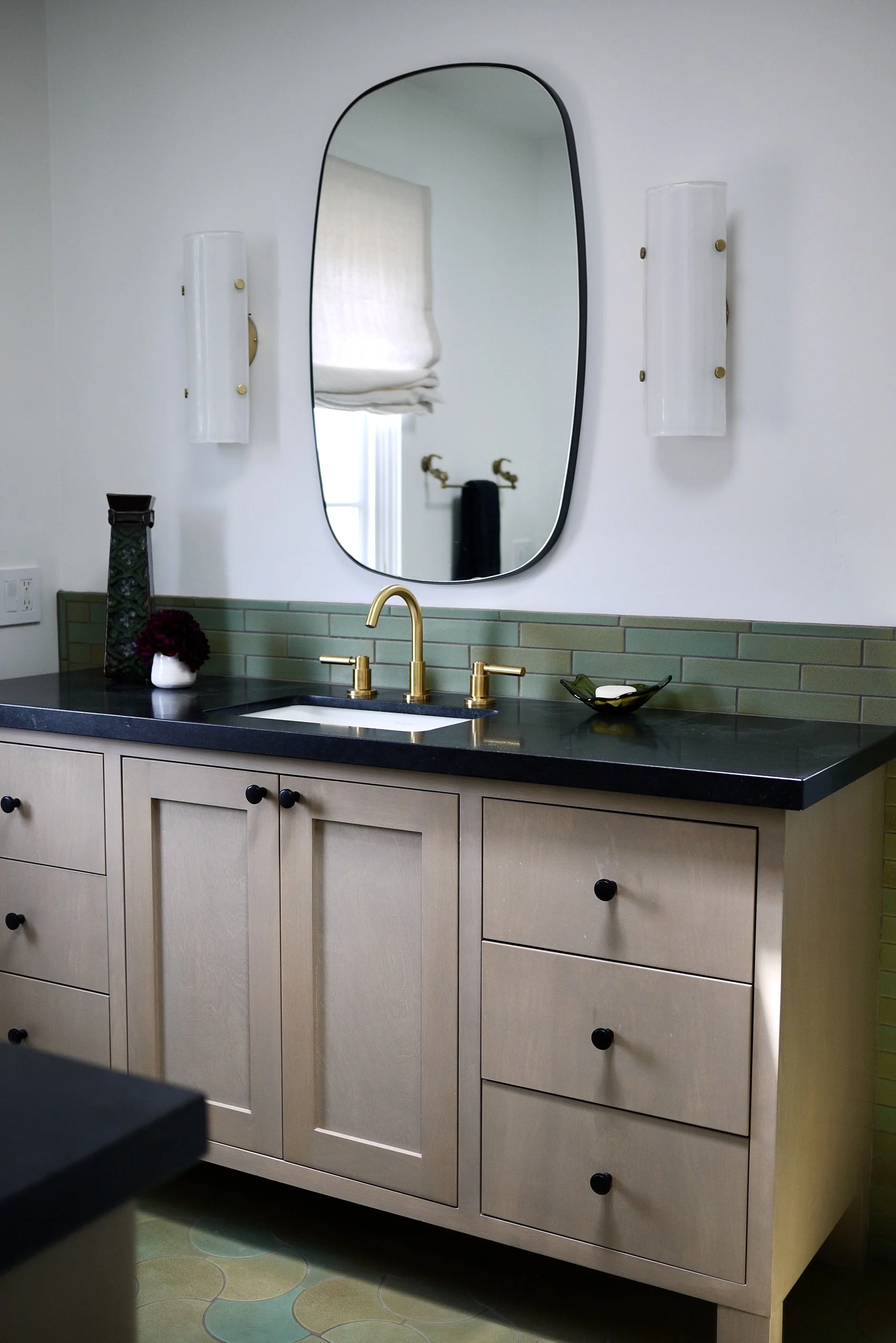 Modern bathroom vanity with wood cabinets, a black countertop, gold faucet, rectangular mirror, and green tile backsplash.