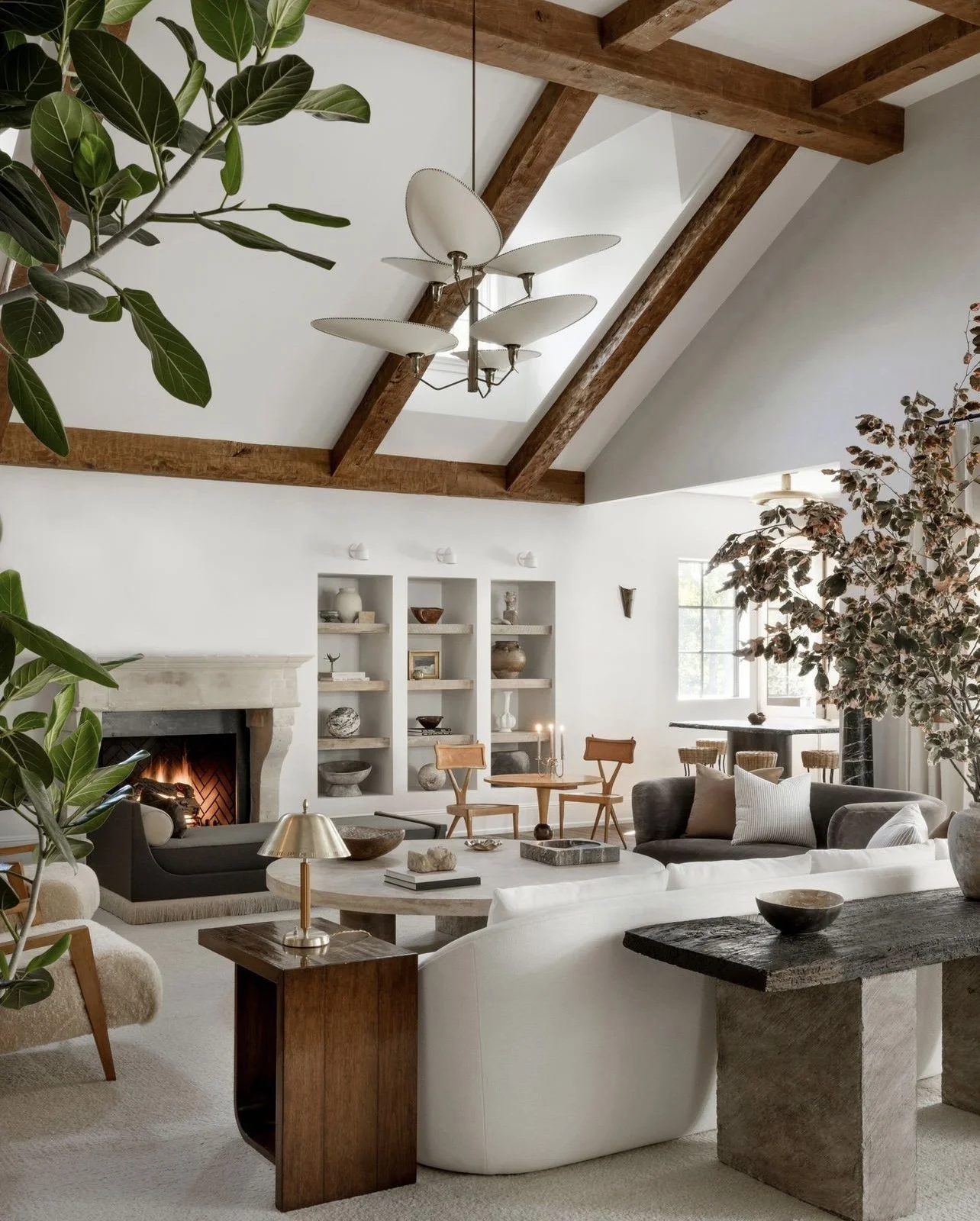 A contemporary living room with vaulted ceiling and exposed wooden beams, featuring neutral-colored furniture, a fireplace, bookshelves, large indoor plants, and modern decor elements.