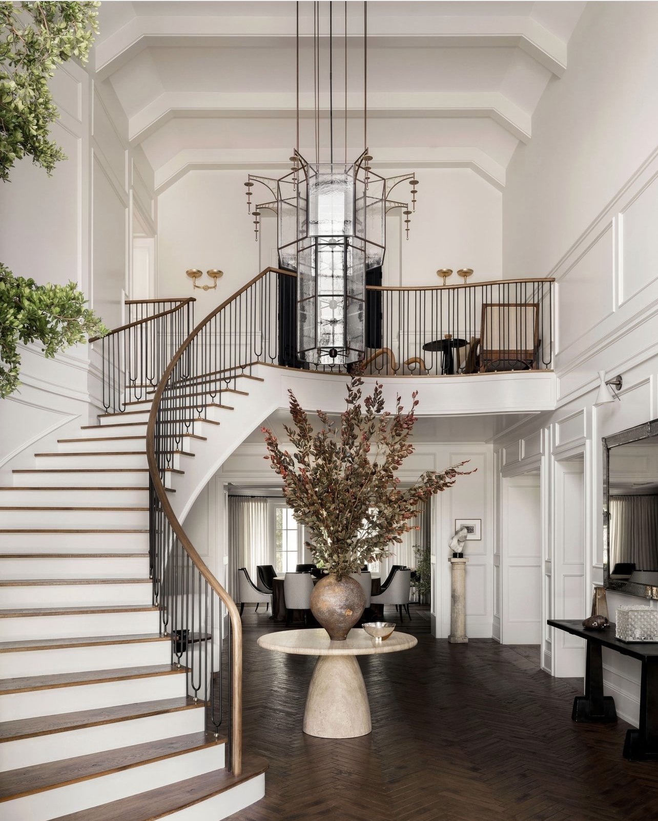 Elegant foyer with a grand spiral staircase, large sculptural chandelier, minimalist round table with decorative vase, and sophisticated decor.
