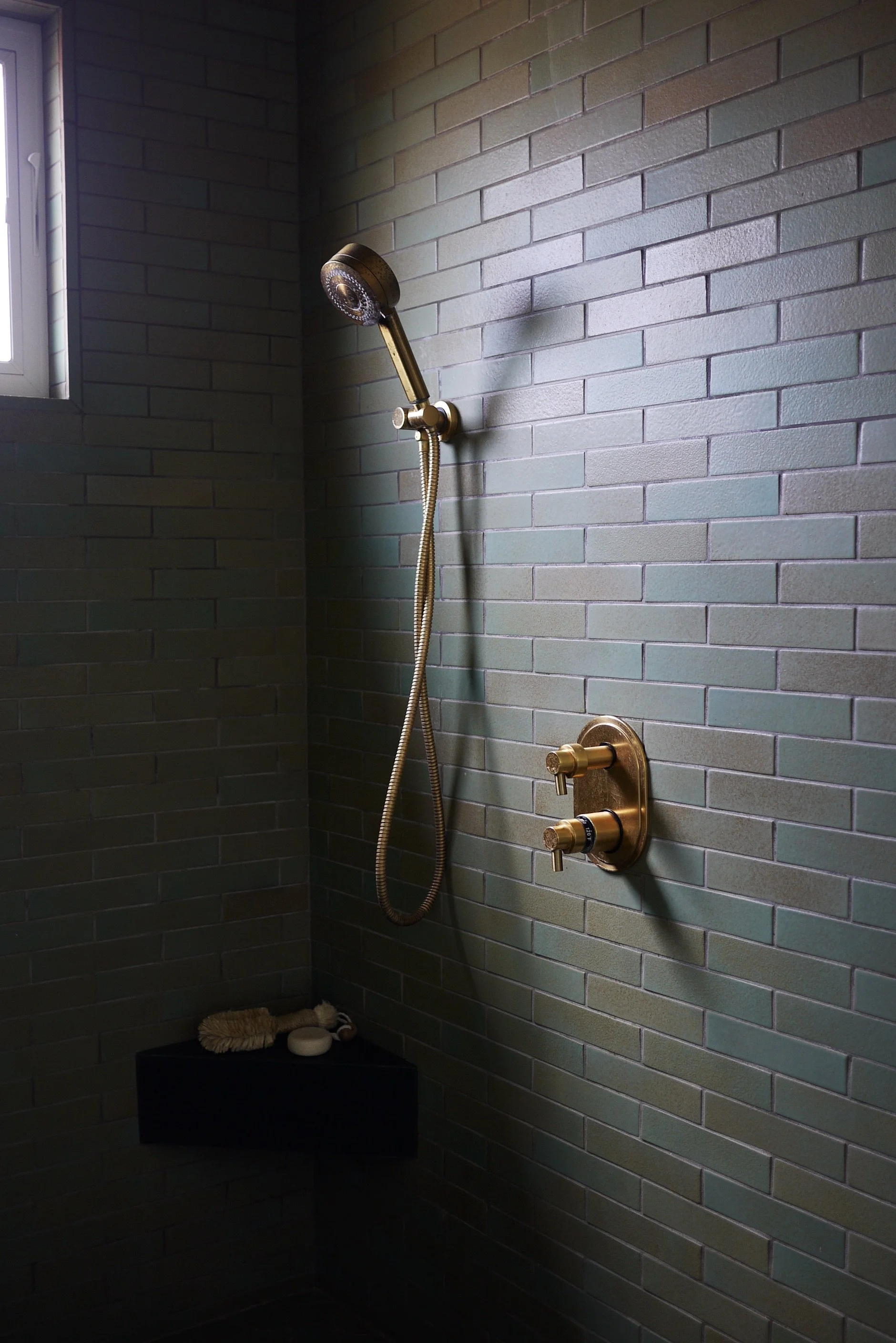 Modern shower with brass fixtures and green tile wall, featuring a corner shelf with a brush and soap.