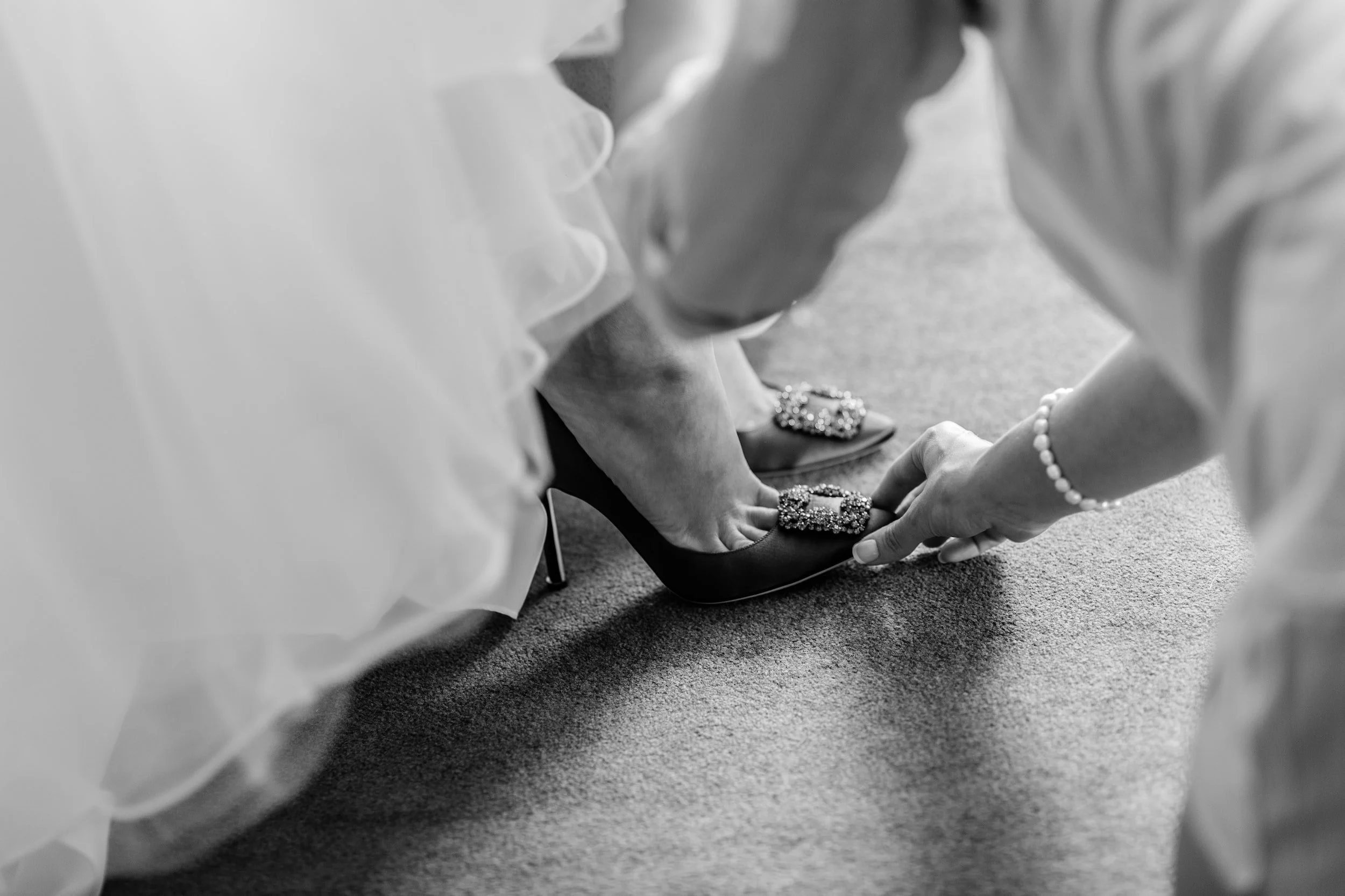 Black and white photo of a person helping someone put on embellished high heels over carpet.