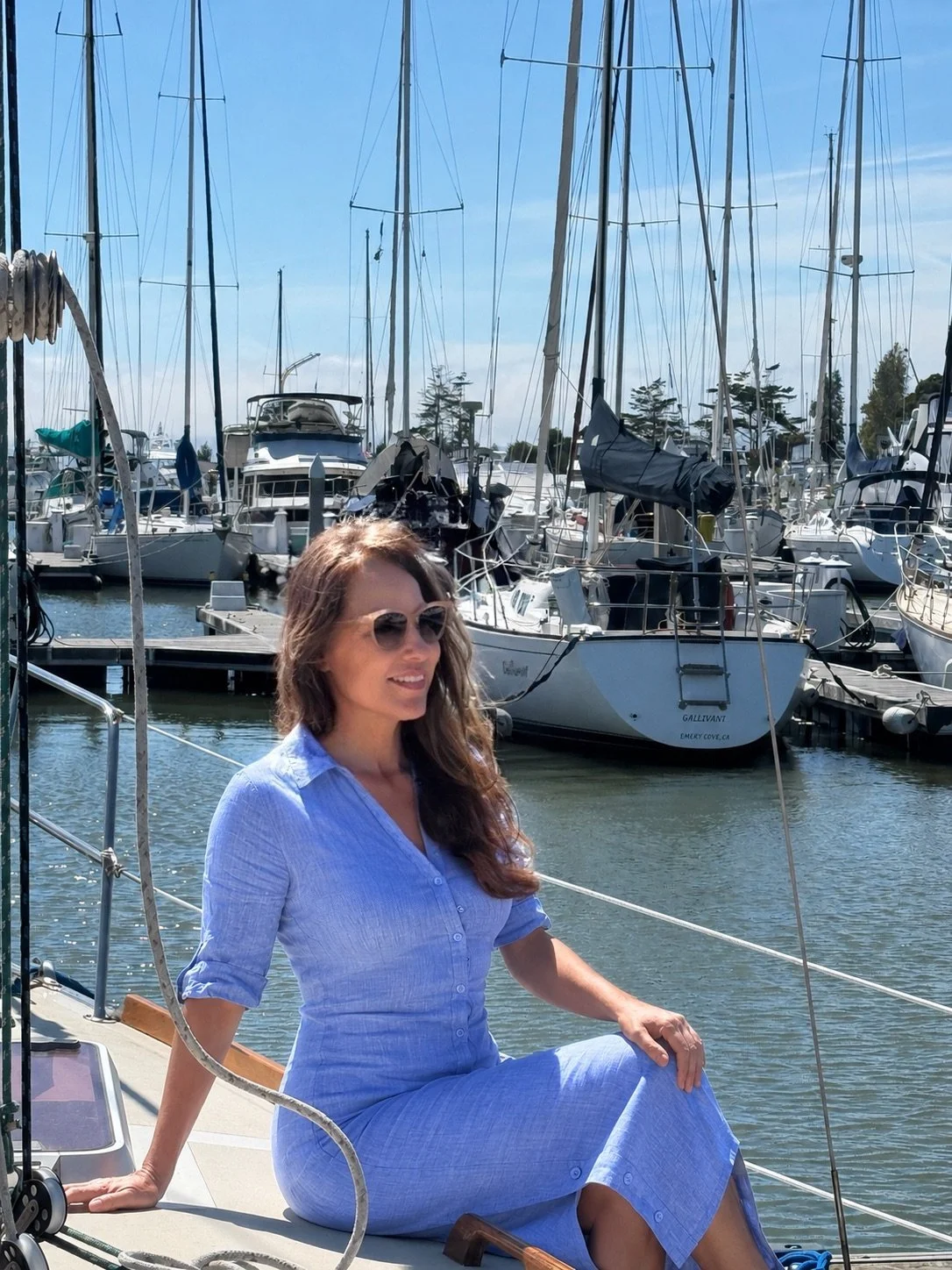 A woman with long hair wearing sunglasses and a light blue dress sitting on a boat docked at a marina filled with sailboats and yachts.