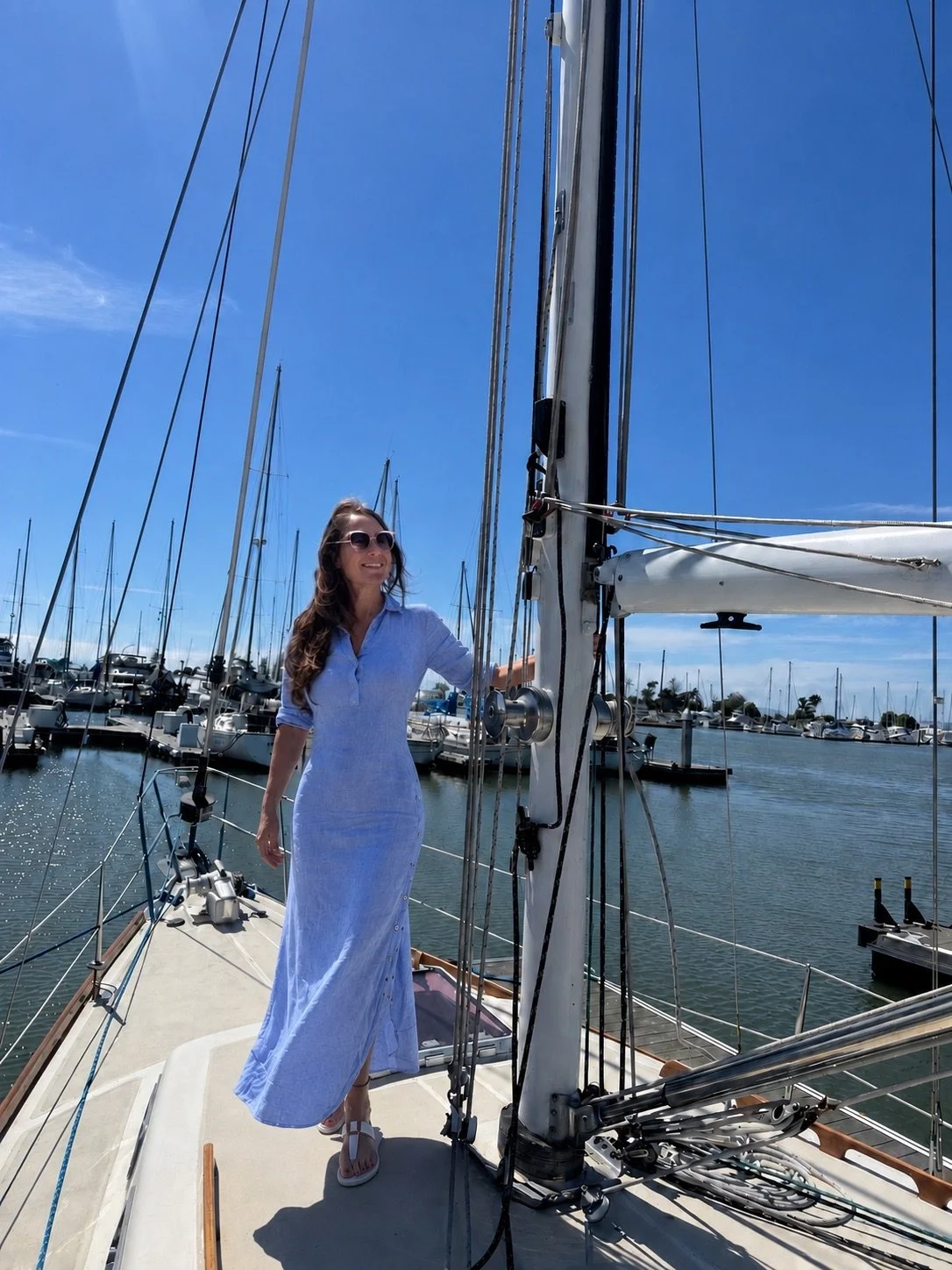 A woman in a light blue dress with sunglasses standing on a sailboat docked at a marina, with other boats and blue sky in the background.