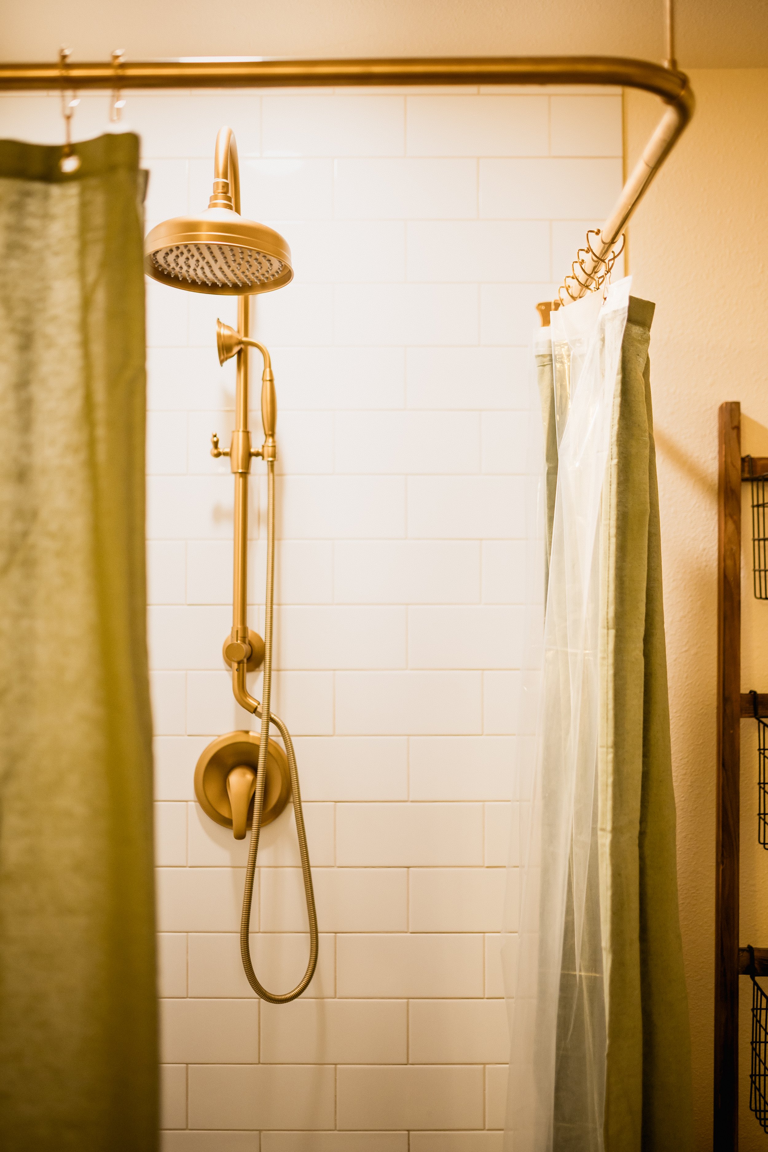 A shower with a gold-colored showerhead, a shower hose, and a control knob, partially enclosed by a green shower curtain on the right and a beige curtain on the left. The background features a white tiled wall and part of a wooden shelf is visible on