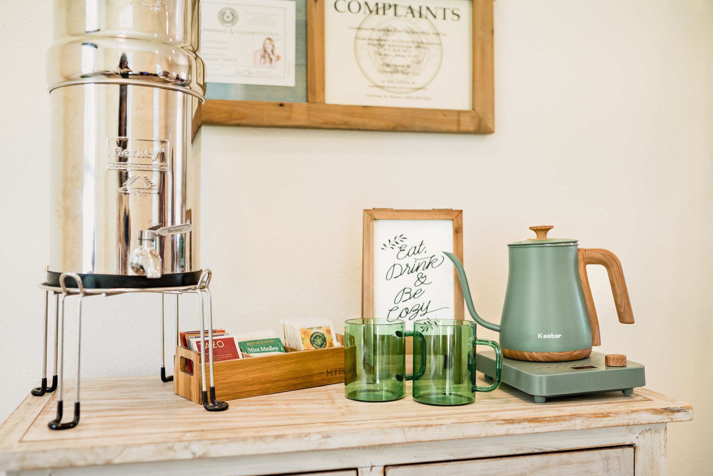 A coffee station with a silver beverage dispenser, green glass mugs, a Keebar coffee maker, and a framed sign that reads 'Eat, Drink & Be Cozy' on a wooden table.