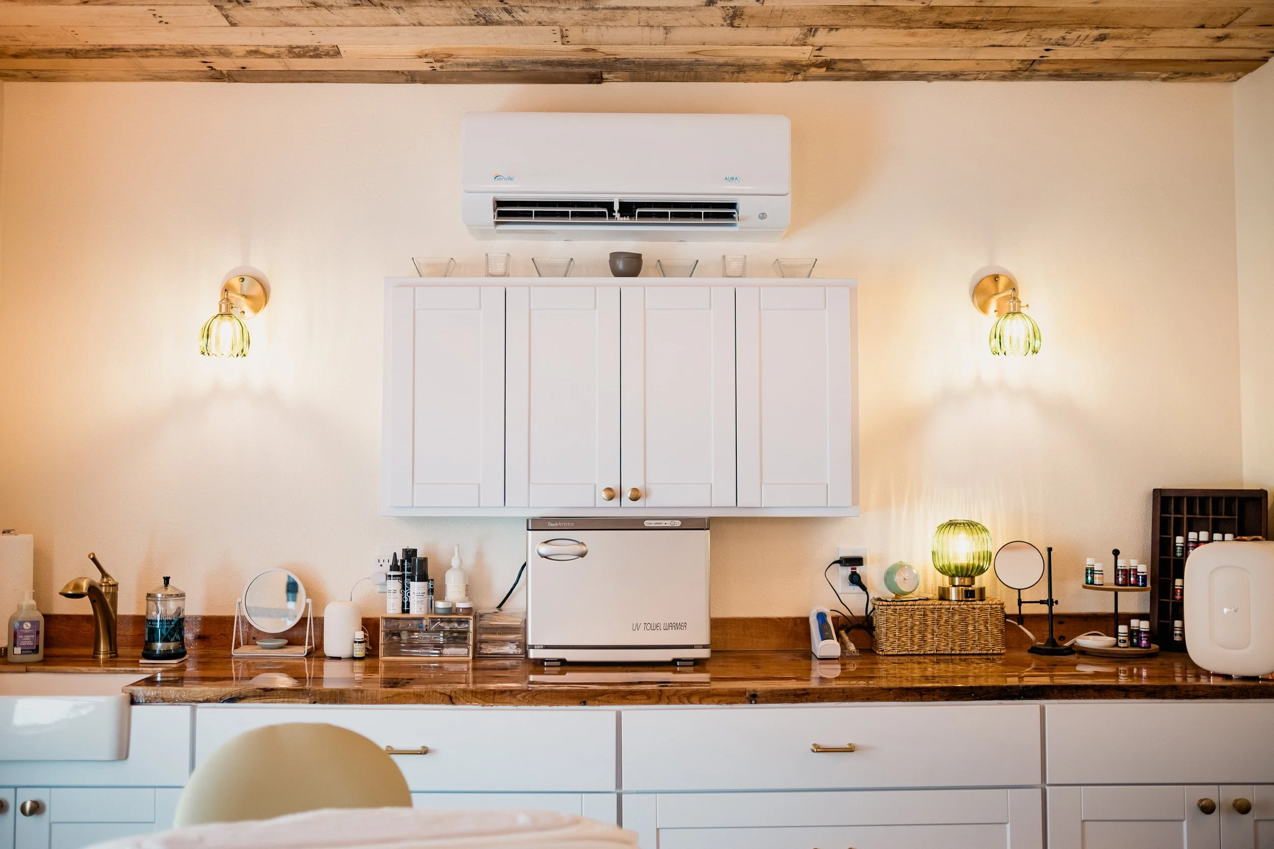 Kitchen with white cabinets, wooden countertop, and wall-mounted air conditioner. Decor includes small bowls, bottles, a lamp, and a mirror on the countertop. Warm lighting.