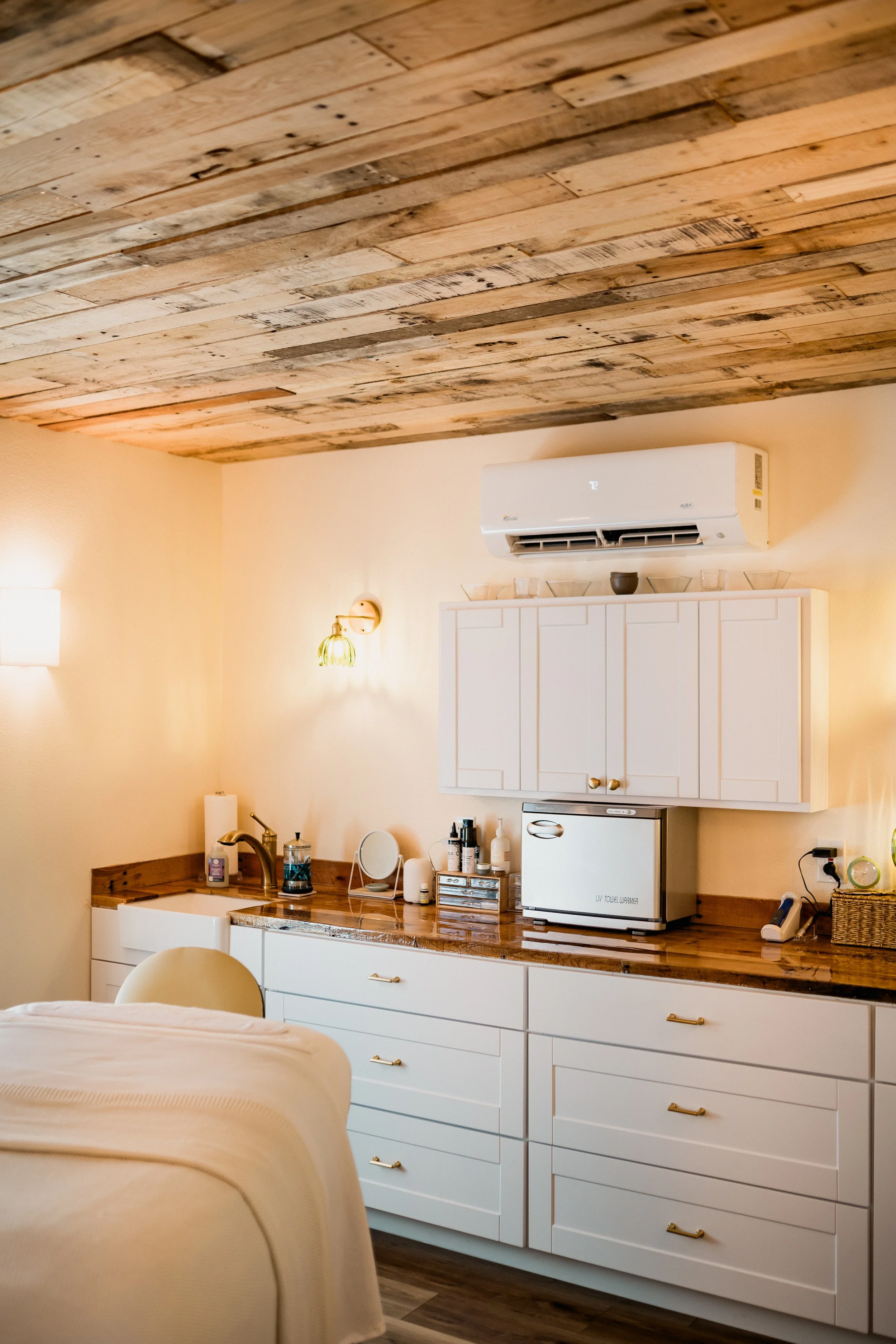 A cozy bedroom with a wooden ceiling, a white cabinet, and a small kitchen area with a countertop, mini fridge, and various items like lamps, bottles, and a mirror.