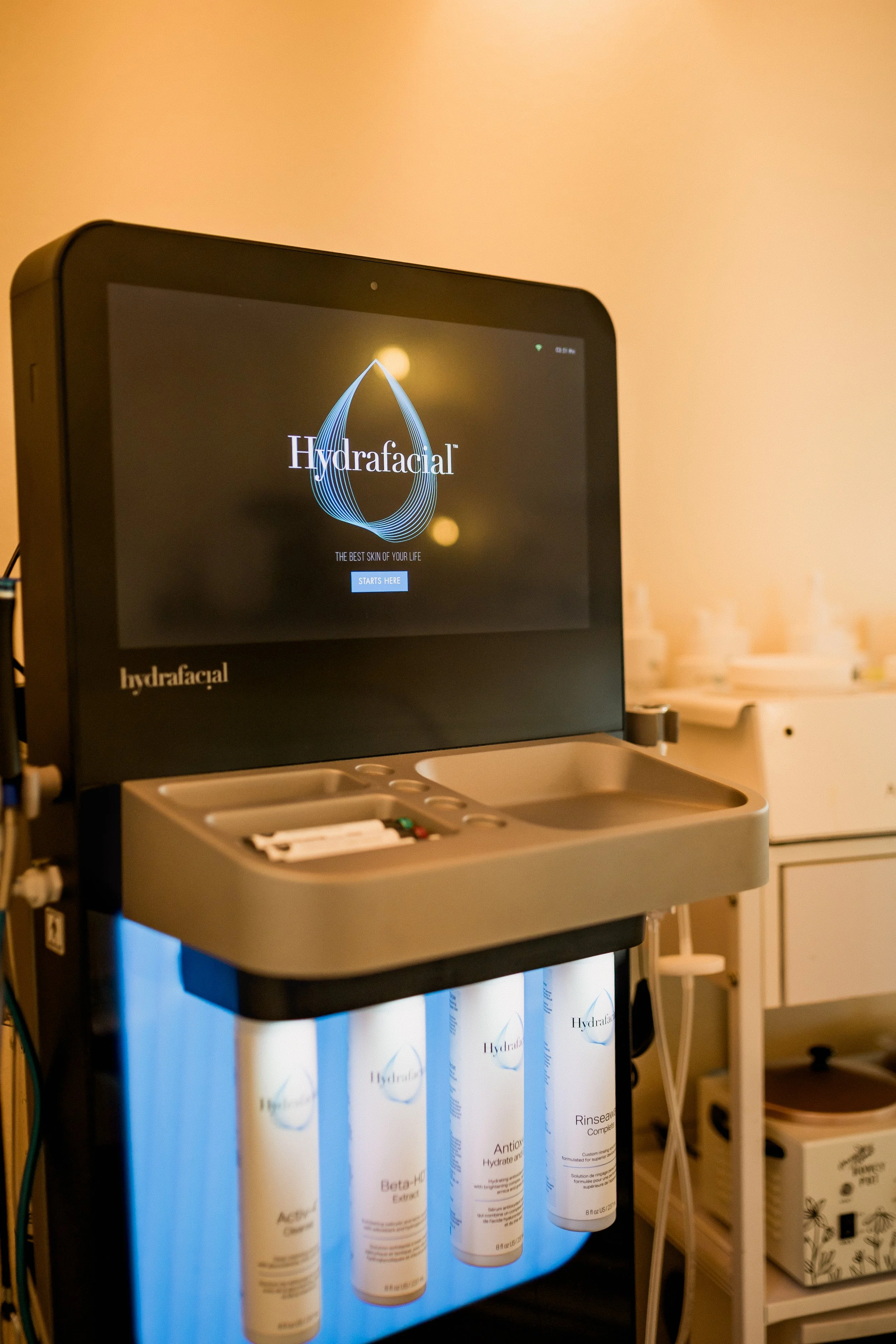 A skincare device with a digital screen displaying the Hydrafacial logo, along with rows of Hydrafacial product bottles underneath, positioned in a clinical or spa setting.