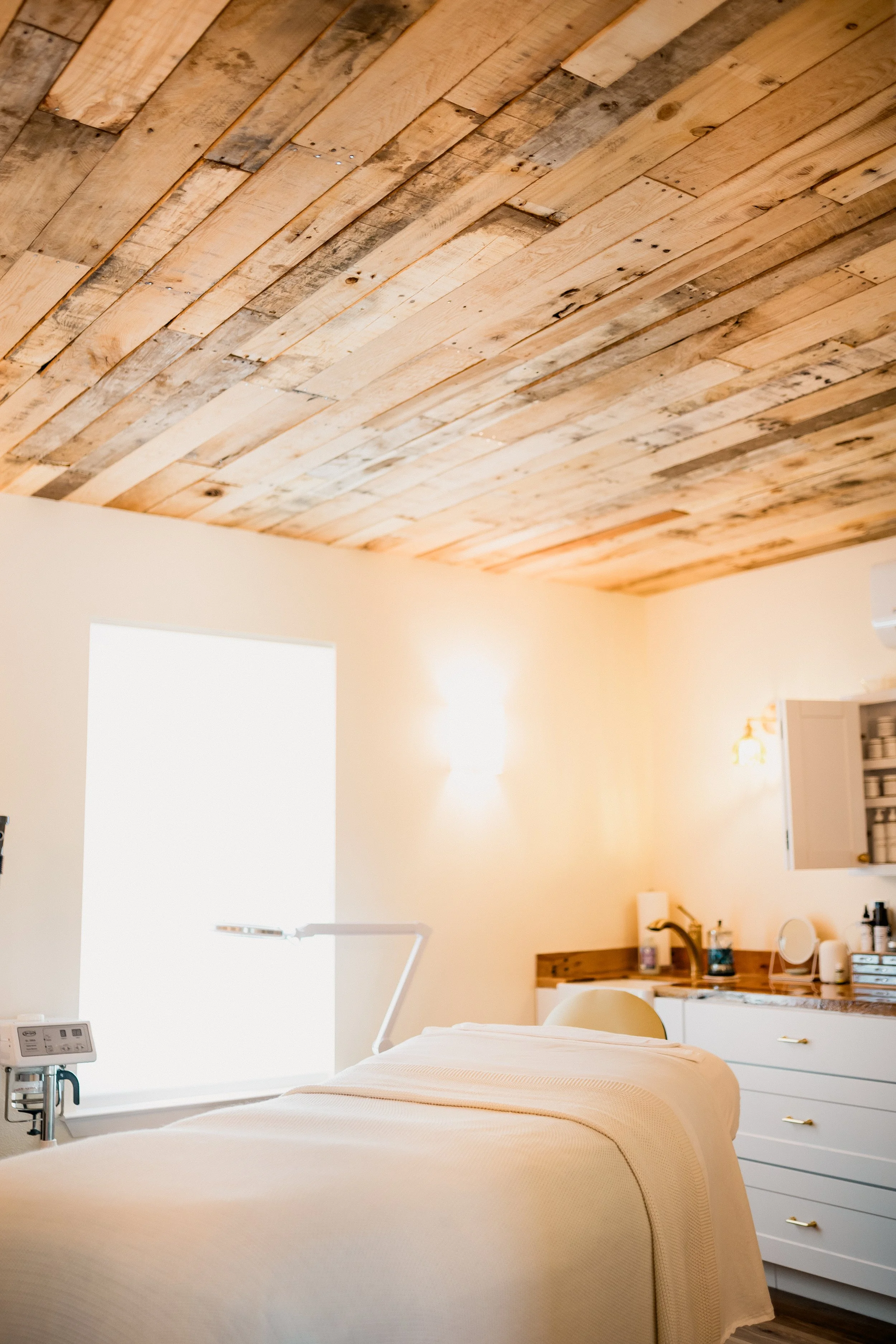 A bedroom with a bed covered with a beige quilt, a wooden headboard, and white drawers. A bedside lamp and various personal items are on the nightstand. The ceiling is made of wooden planks, and there are two large bright windows letting in natural l