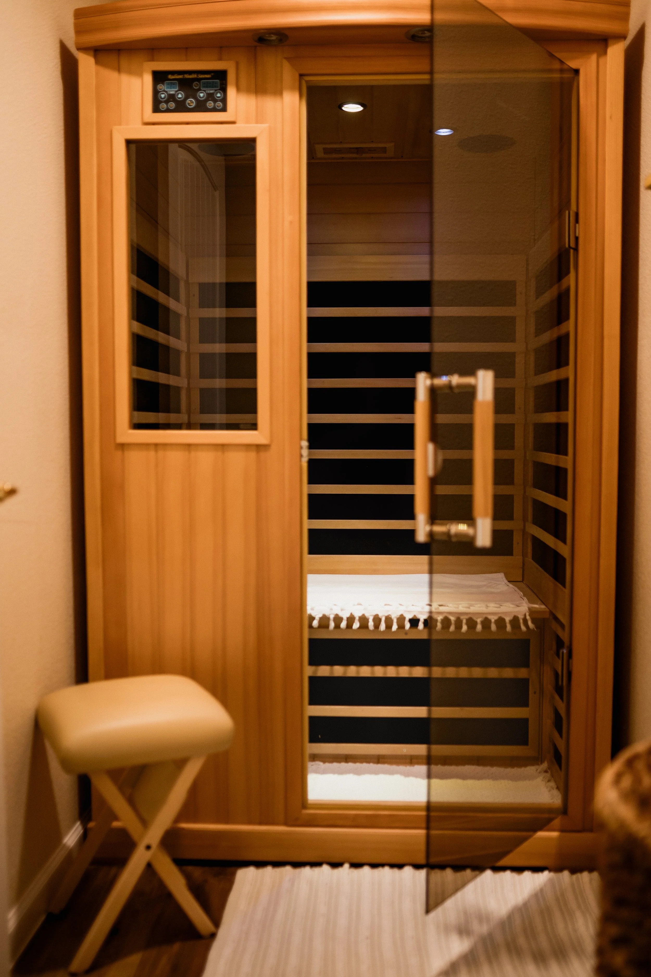 A glass door sauna with a wooden interior reflected in the mirror, with a small cream-colored stool in front and a striped rug on the floor.