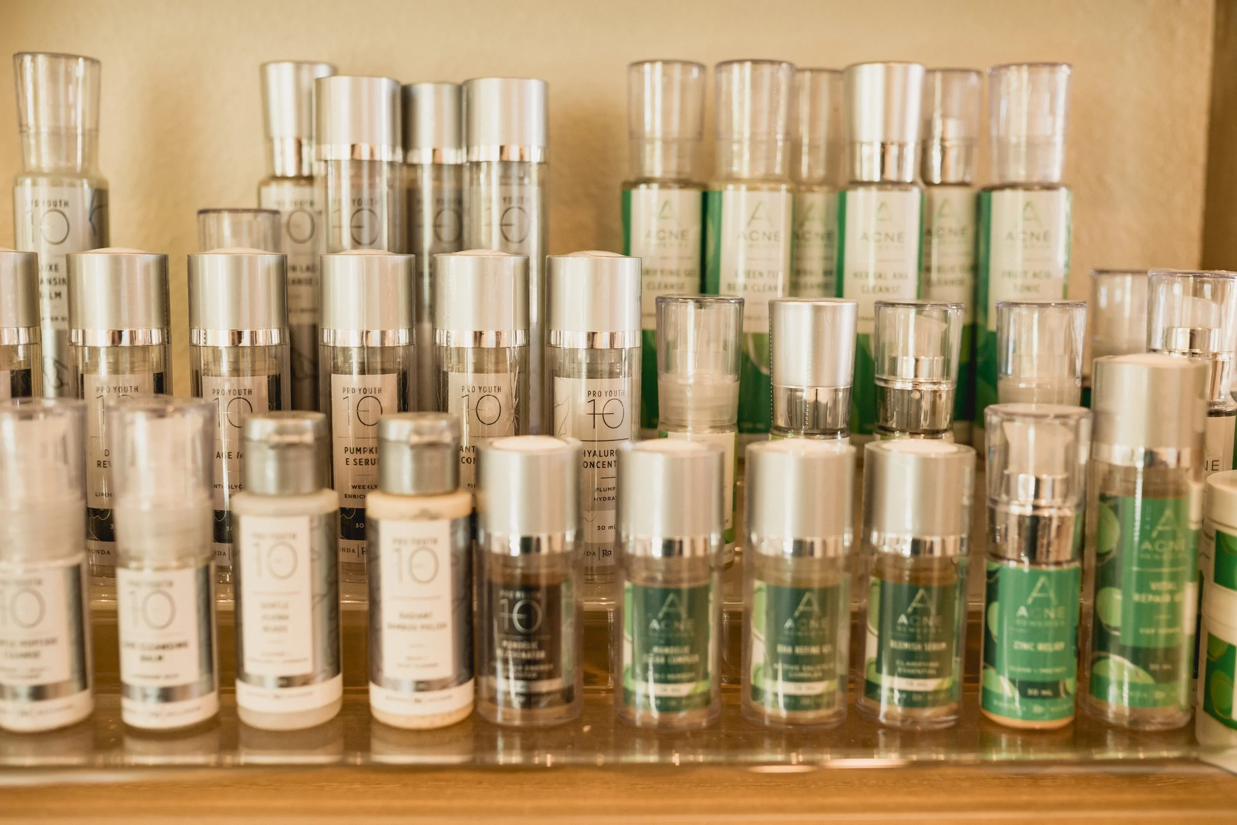 Collection of skincare or cosmetic bottles with white caps and green labels, arranged on a wooden shelf.