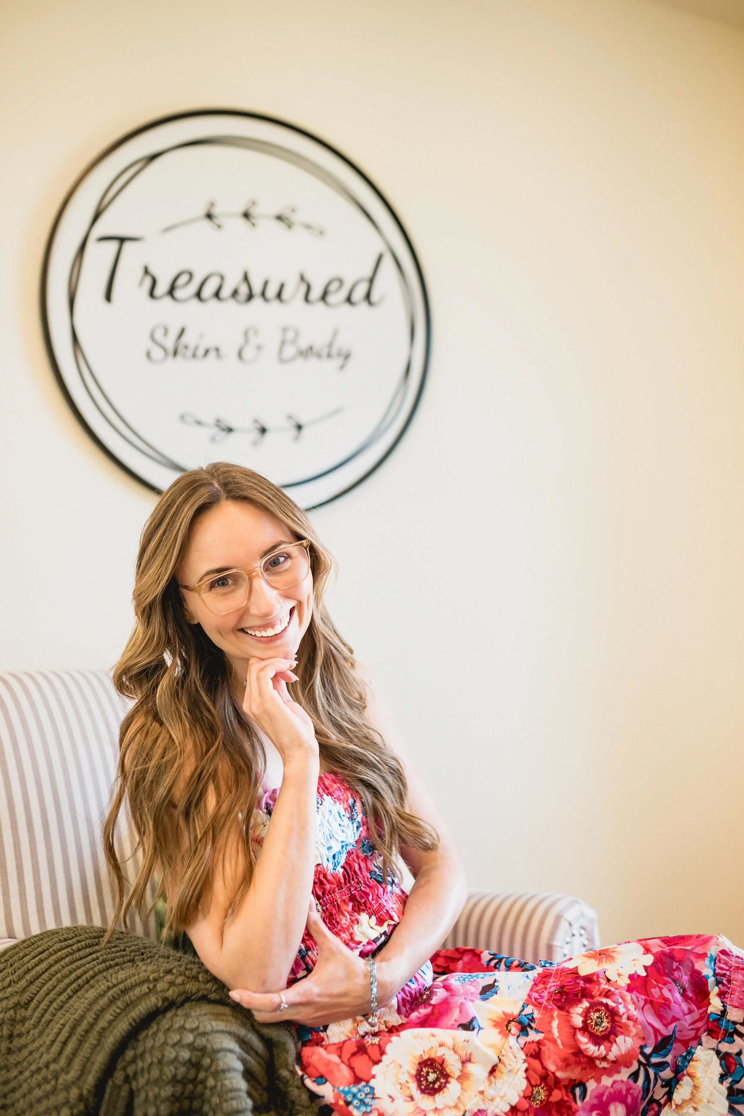 A smiling woman with long wavy hair, glasses, and colorful floral dress sitting on a striped sofa with a brown knit blanket, in front of a wall with a round sign that reads 'Treasured Skin & Body' decorated with a botanical illustration.