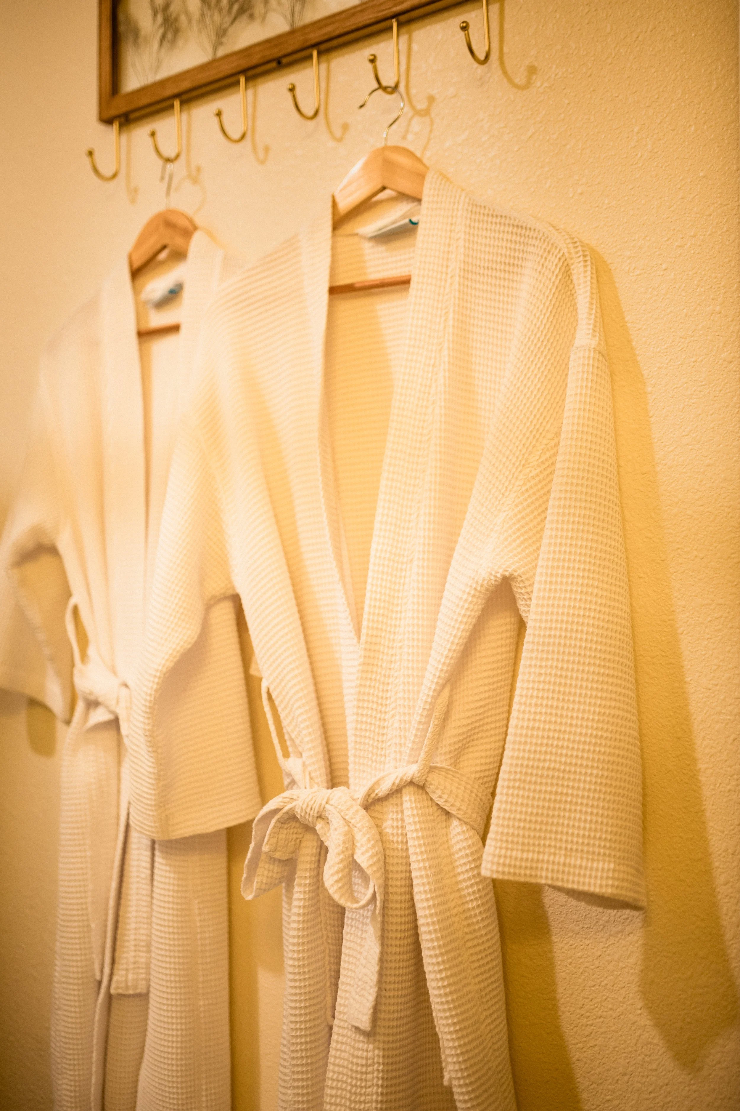 Two cream-colored waffle-knit robes with belts hanging on hooks in a bathroom or spa setting.