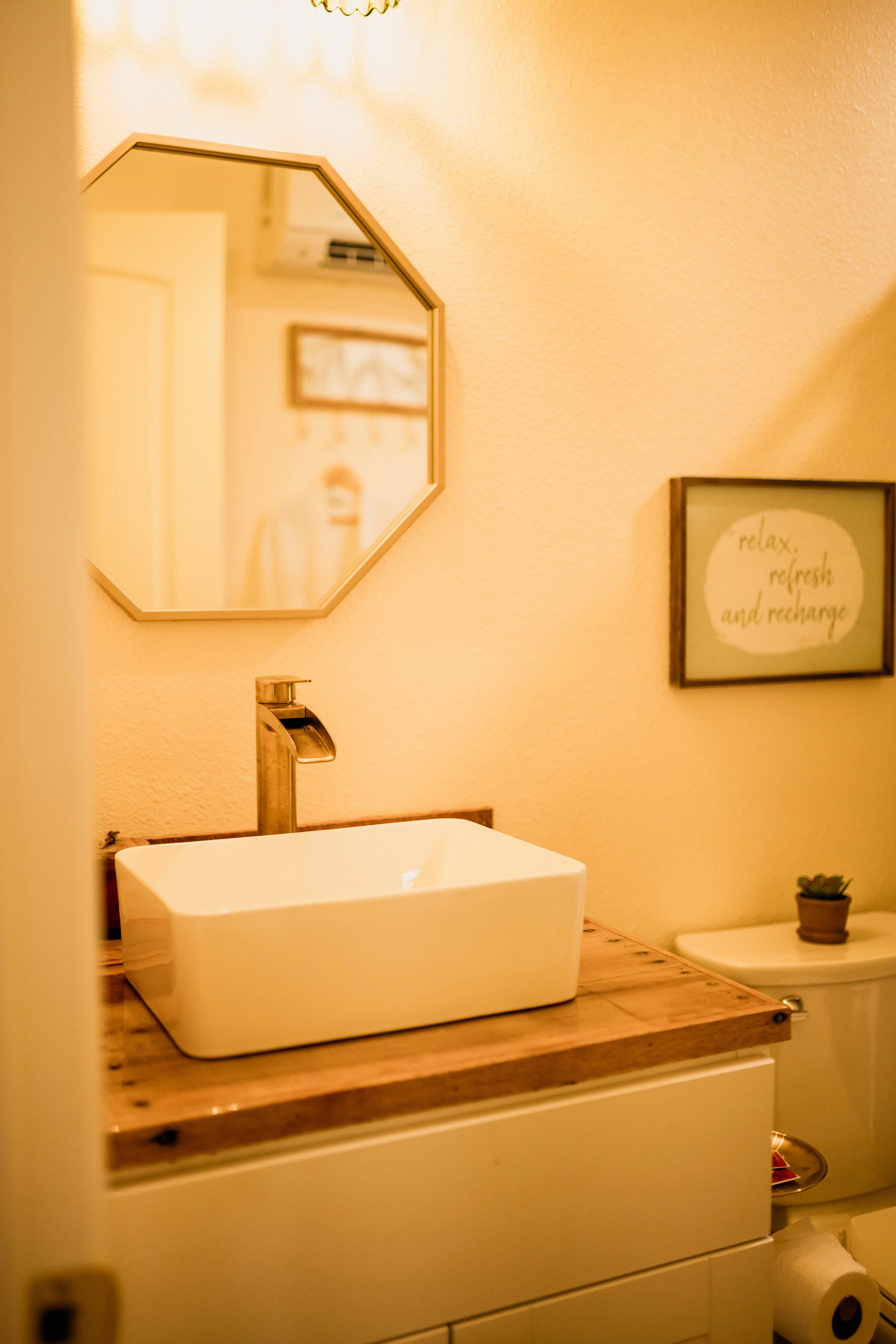 A bathroom with a white rectangular sink on a wooden vanity, a gold-colored faucet, a small potted succulent, a mirror, and wall decor including a frame with the words "relax, refresh, and recharge."