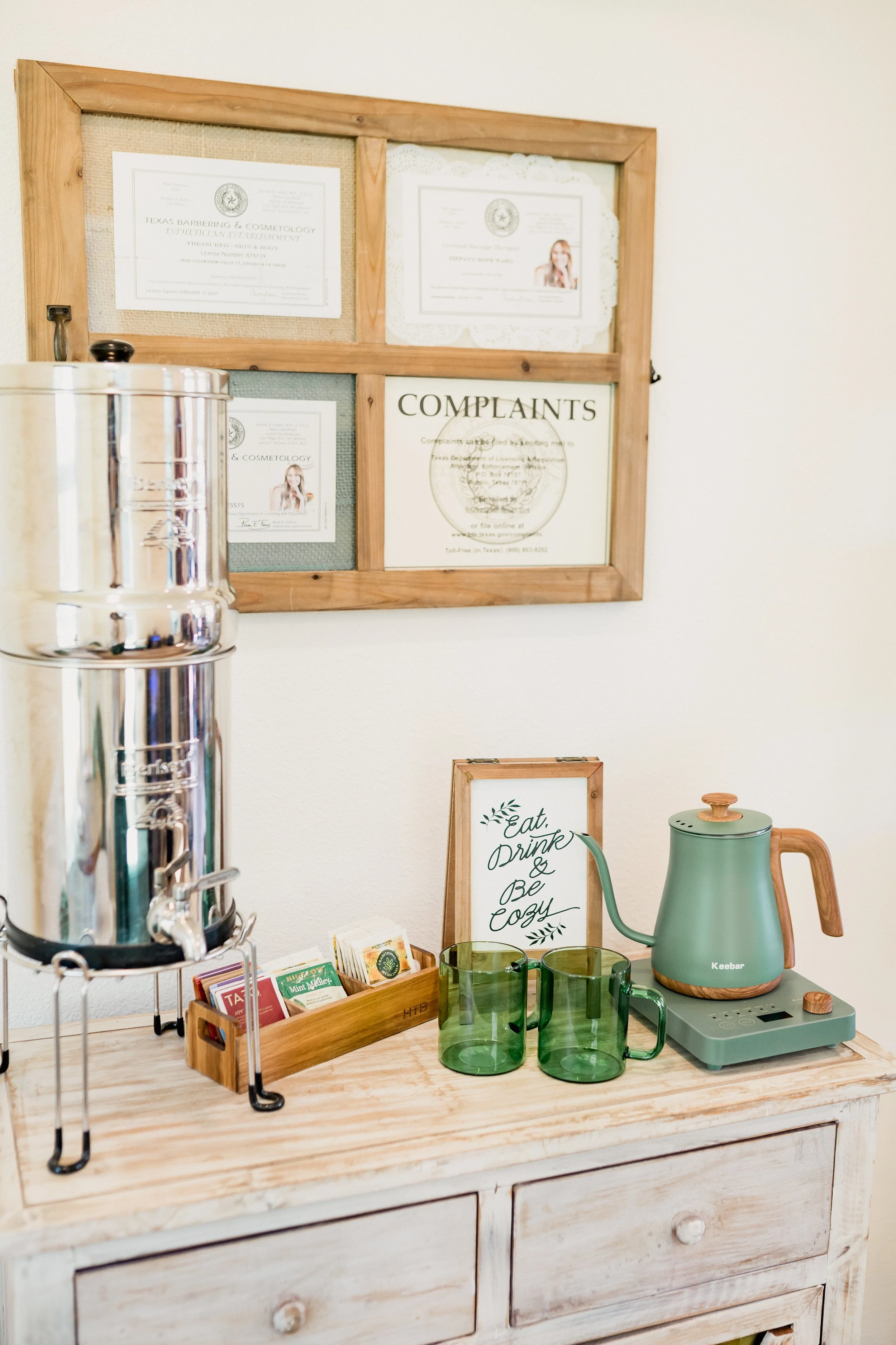A wooden dresser with a tea kettle, glass mugs, herbal tea packets, and a sign that reads 'Eat, Drink & Be Cozy'. Behind it, a wall has framed certificates and a complaint form.