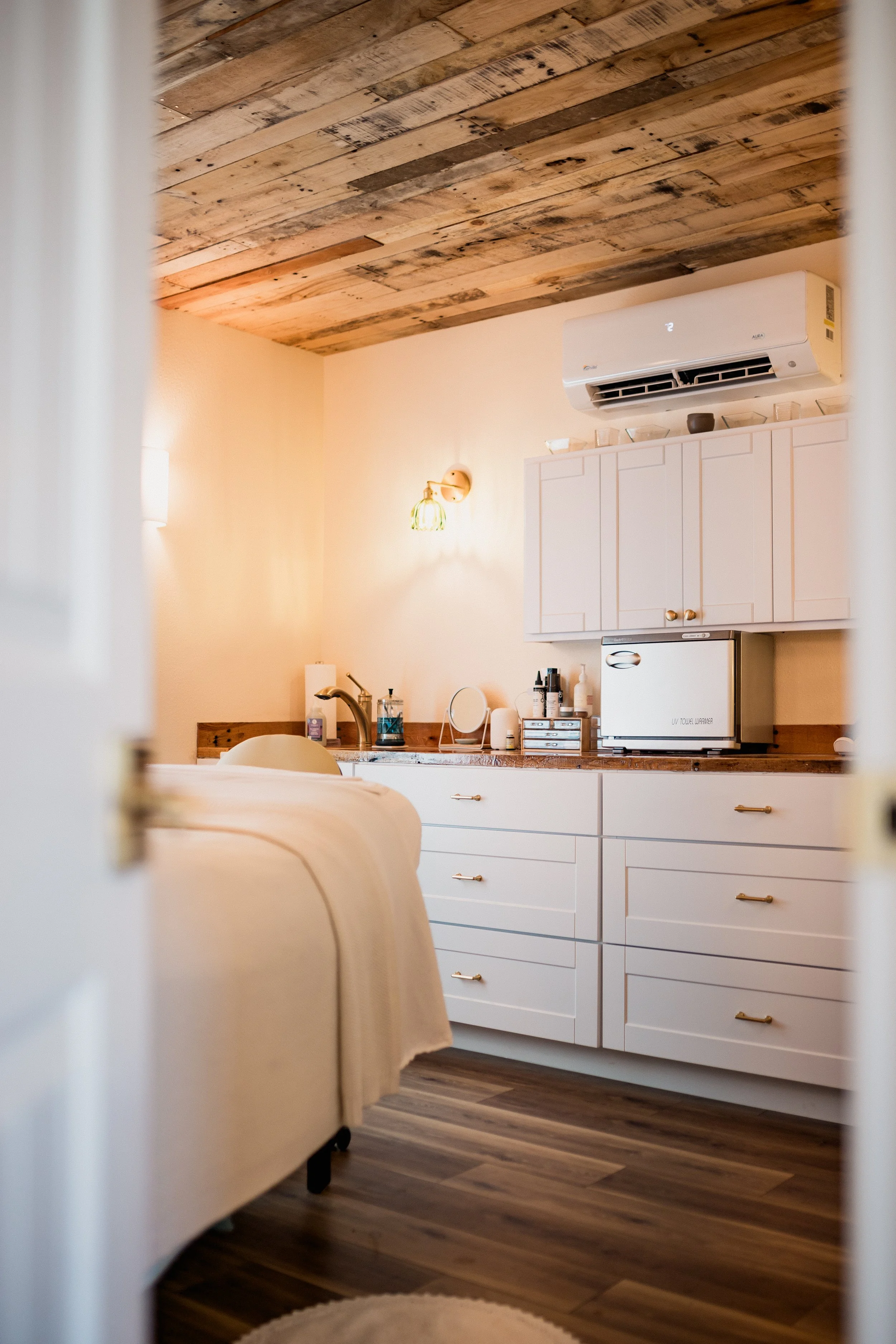 The image shows a cozy kitchen with a wooden ceiling, a white cabinet, a small refrigerator, and various kitchen essentials on the countertop. The photo is taken from behind a partially opened door or curtain, revealing a warm and inviting interior.