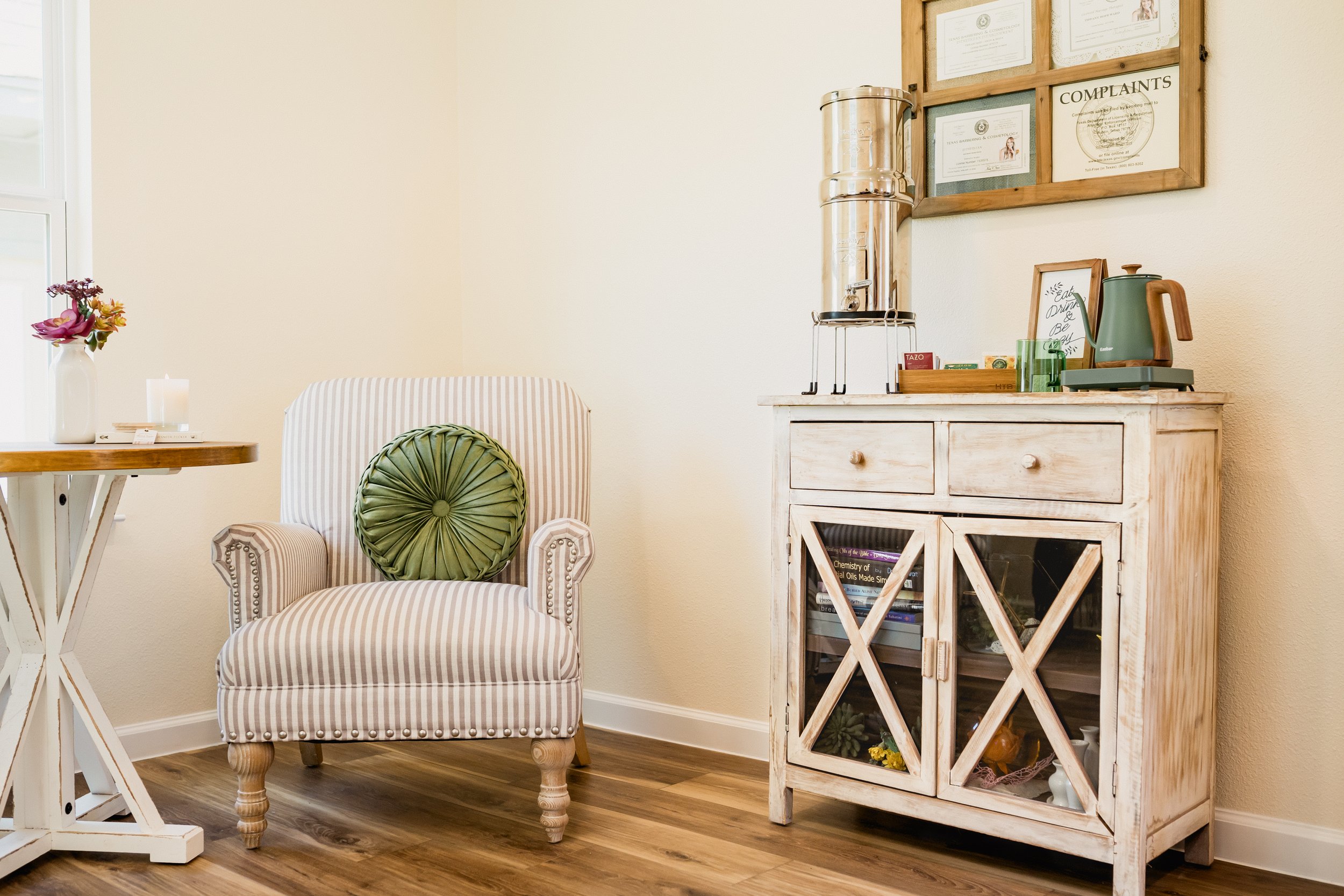 A cozy sitting area with a striped armchair featuring a green round pillow. To the left, a small white table with a vase of flowers and a lit candle. To the right, a rustic white wooden cabinet with glass doors, containing books and decorative items,
