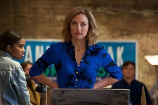 Lola Glaudini in "Ray Donovan" wearing a blue shirt standing with her hands on her hips in a room with brick walls and people in the background.
