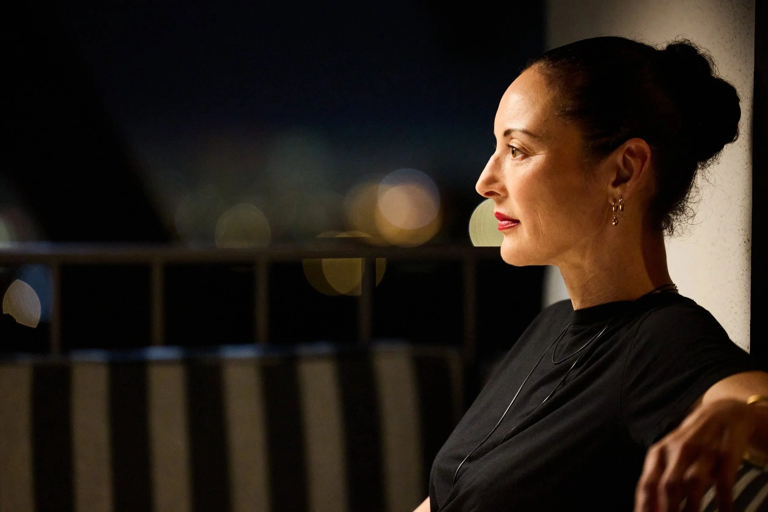 Lola Glaudini in short film "Tu Viens D'où." Lola's profile with dark hair in a bun, wearing earrings, looking to the right, with a blurred night cityscape background.
