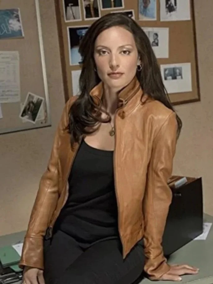 Lola Glaudini. Elle Greenaway in Criminal Minds. With long dark hair, wearing a tan leather jacket over a black top, sitting on a desk in an office or classroom setting with bulletin boards and photos in the background.