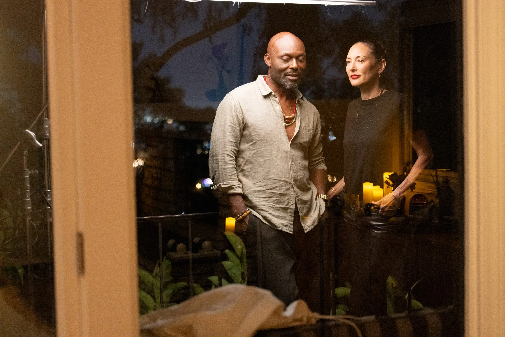 Lola Glaudini and Jimmy Jean-Louis in "Tu Viens D'où" - we see them through a window at night, standing on a balcony close together with trees and lights in the background.