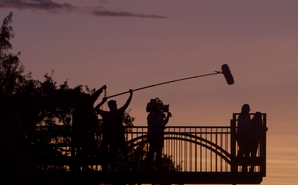 Silhouettes of a film crew filming on a bridge at sunset, one person holding a boom microphone, another operating a camera, and two individuals standing together. Behind the scenes on short film "Mighty" by Lola Glaudini.