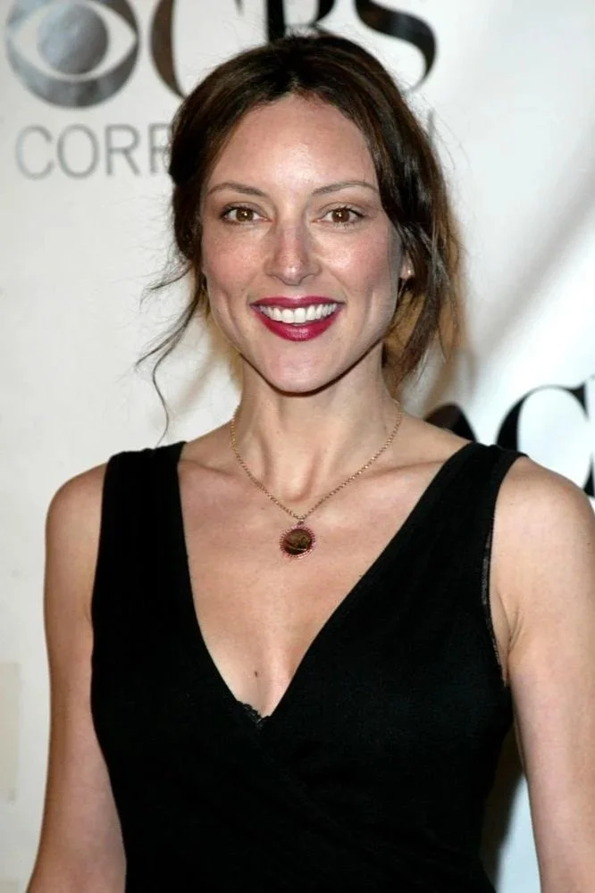 Actress Lola Glaudini wearing a black sleeveless dress and a necklace with a round pendant, smiling in front of a backdrop with CBS logos.