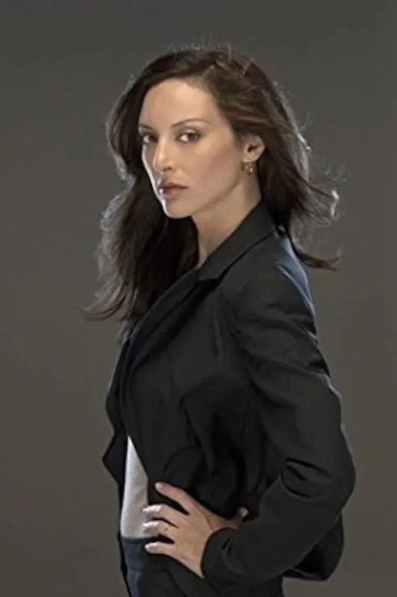 Lola Glaudini as Elle Greenaway from "Criminal Minds." She's got long brown hair and is wearing a black blazer and white top, posing confidently.