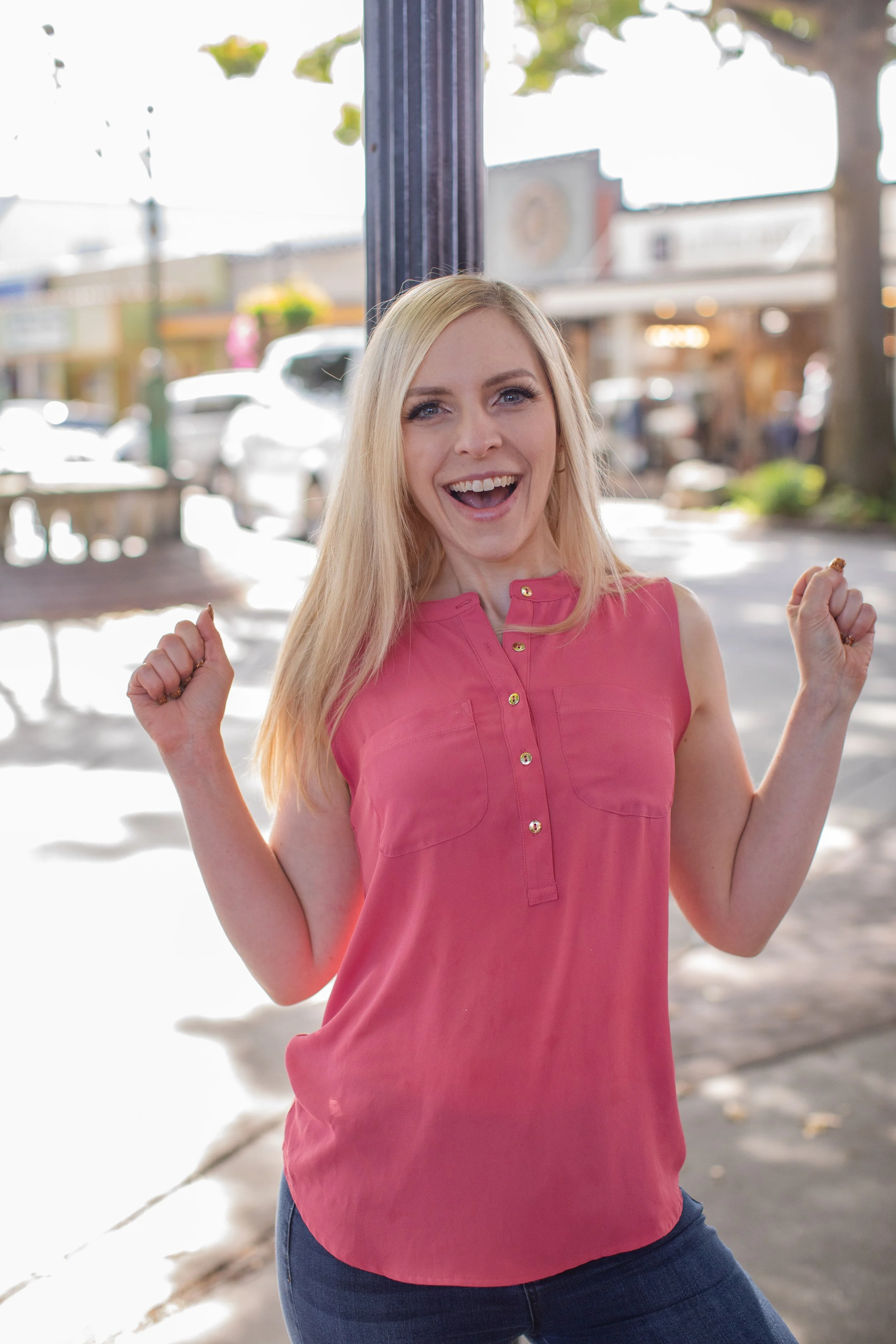 A woman with blonde hair wearing a pink sleeveless top and jeans, celebrating outdoors on a sunny day.