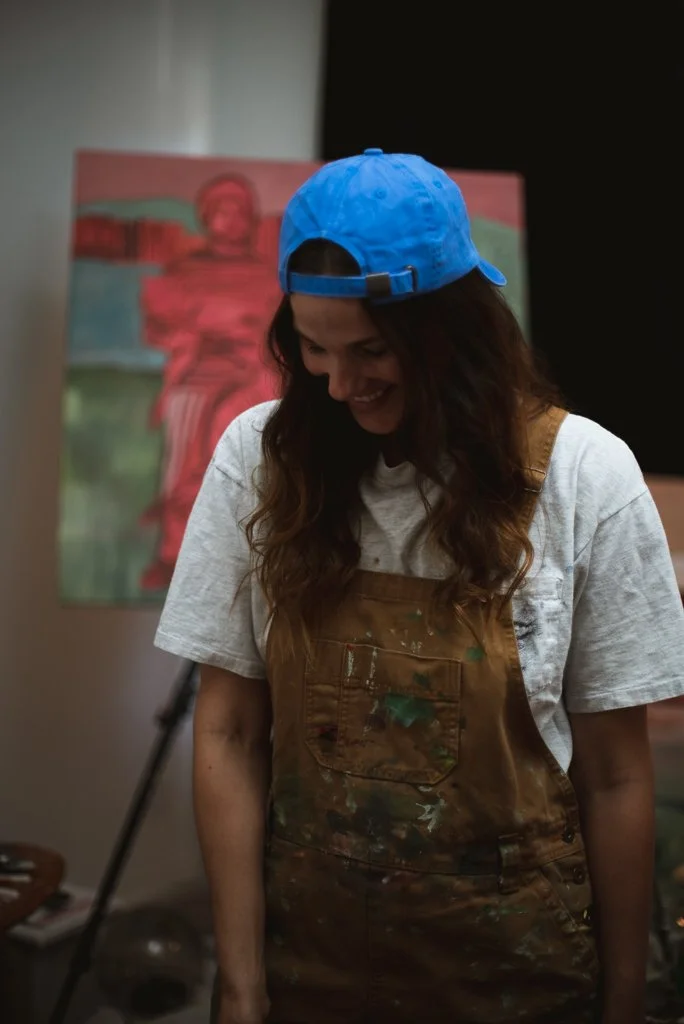 Artist wearing a blue cap and paint-splattered overalls standing in front of a colorful painting on an easel.