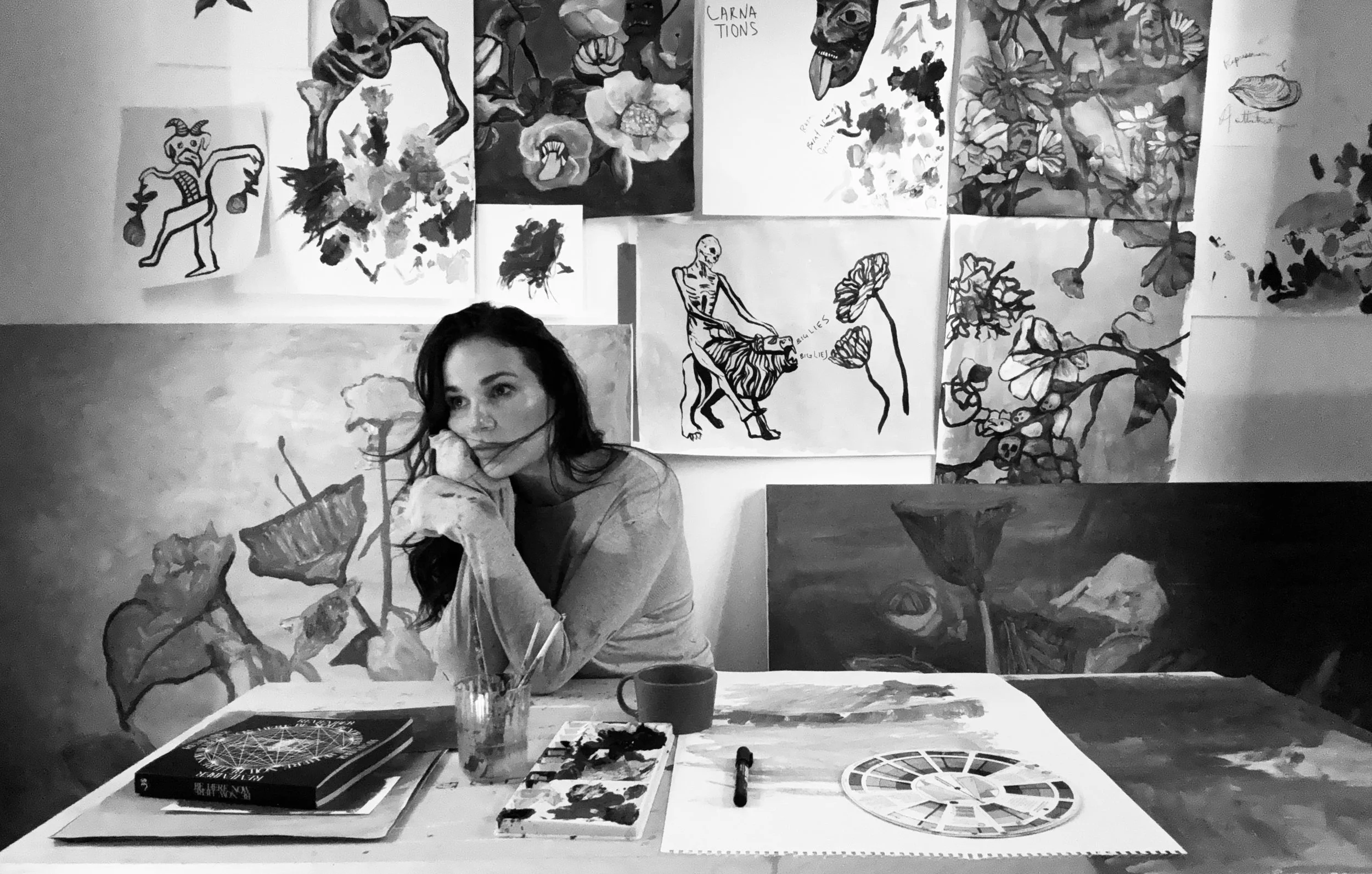 Artist sitting at table in studio surrounded by paintings and sketches, looking contemplative.