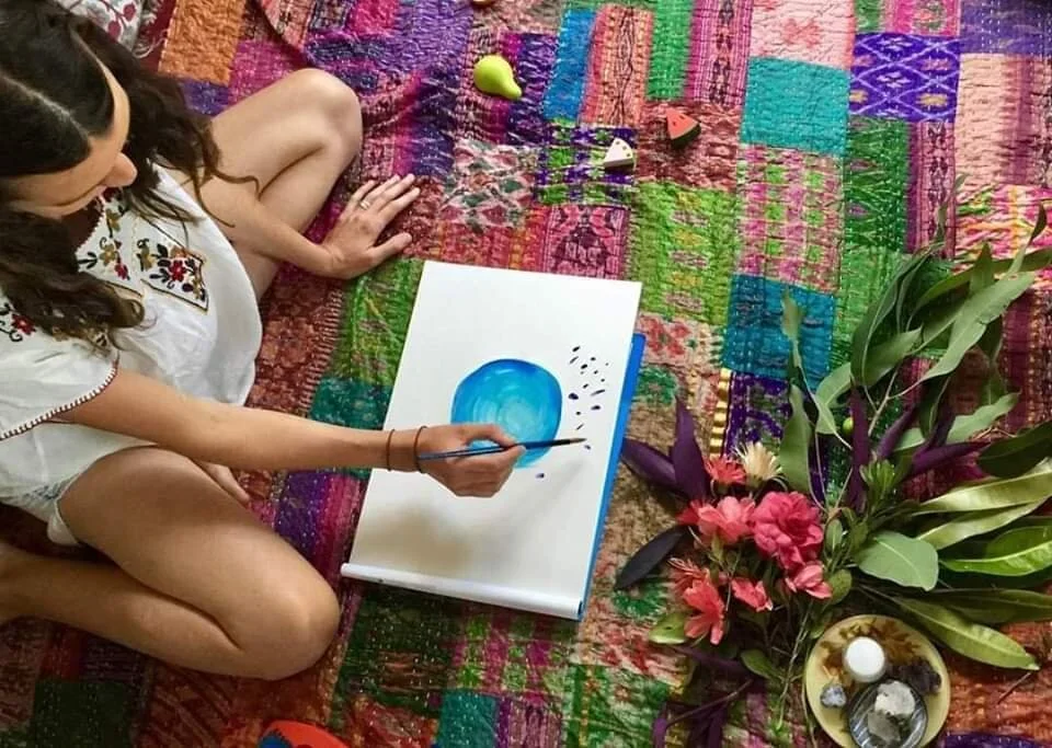 A woman sits on a rug, painting on a canvas with a brush. She is surrounded by various objects, including fruits, a plant, and a bowl with candles, on a patterned surface.