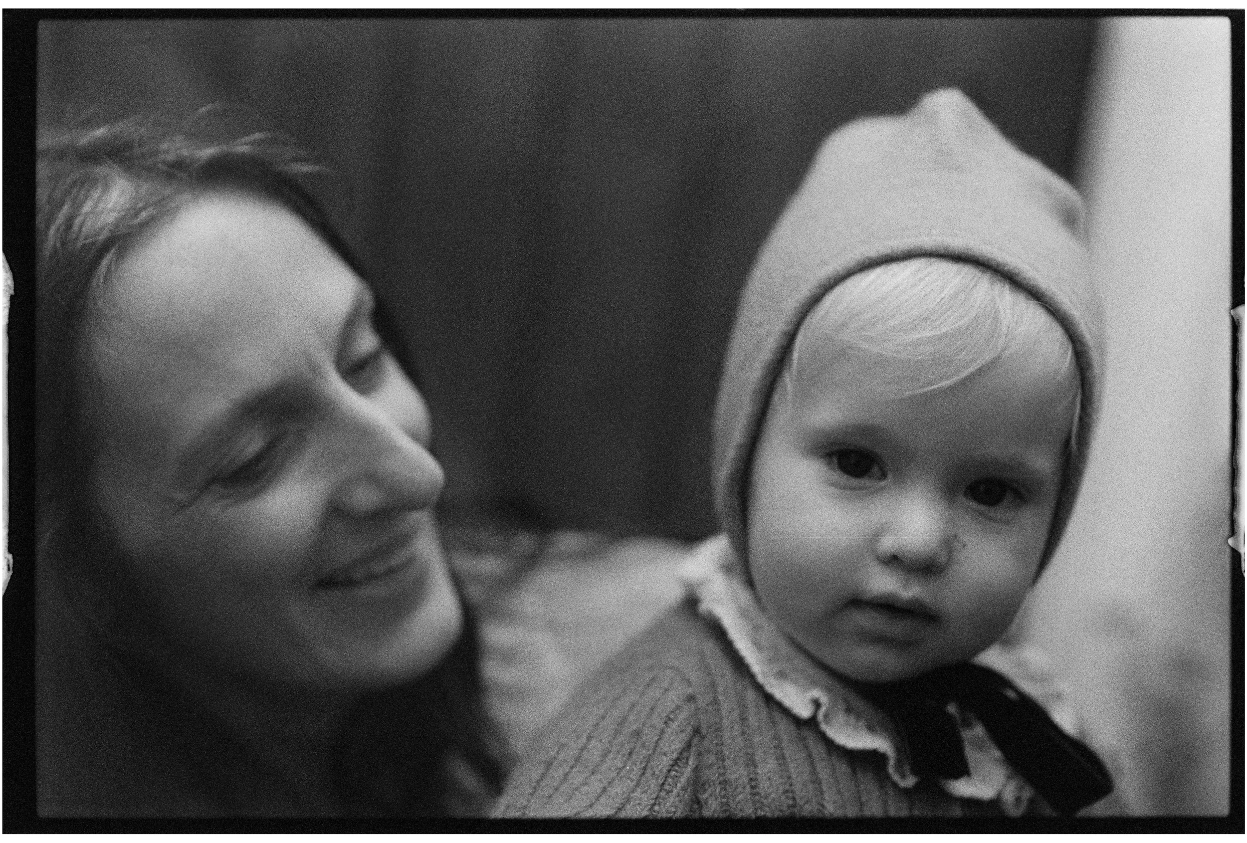 A black-and-white photo of a smiling woman with long hair on the left and a young child with light hair, wearing a beanie hat, on the right.