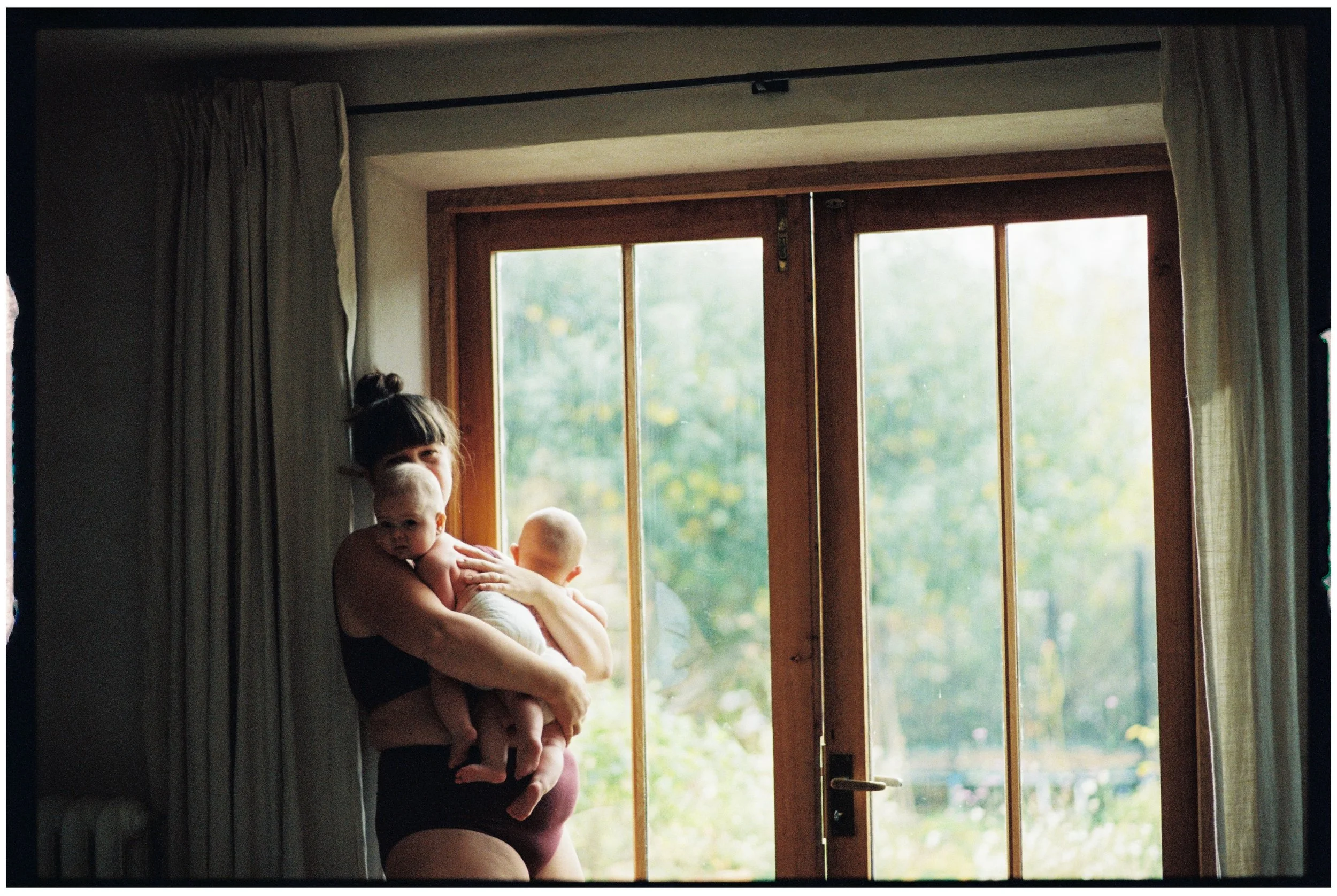 A woman in black underwear holding two young children, one facing forward and the other with back to camera, standing near a large wooden framed glass door with curtains on sides, in a brightly lit room with greenery outside.