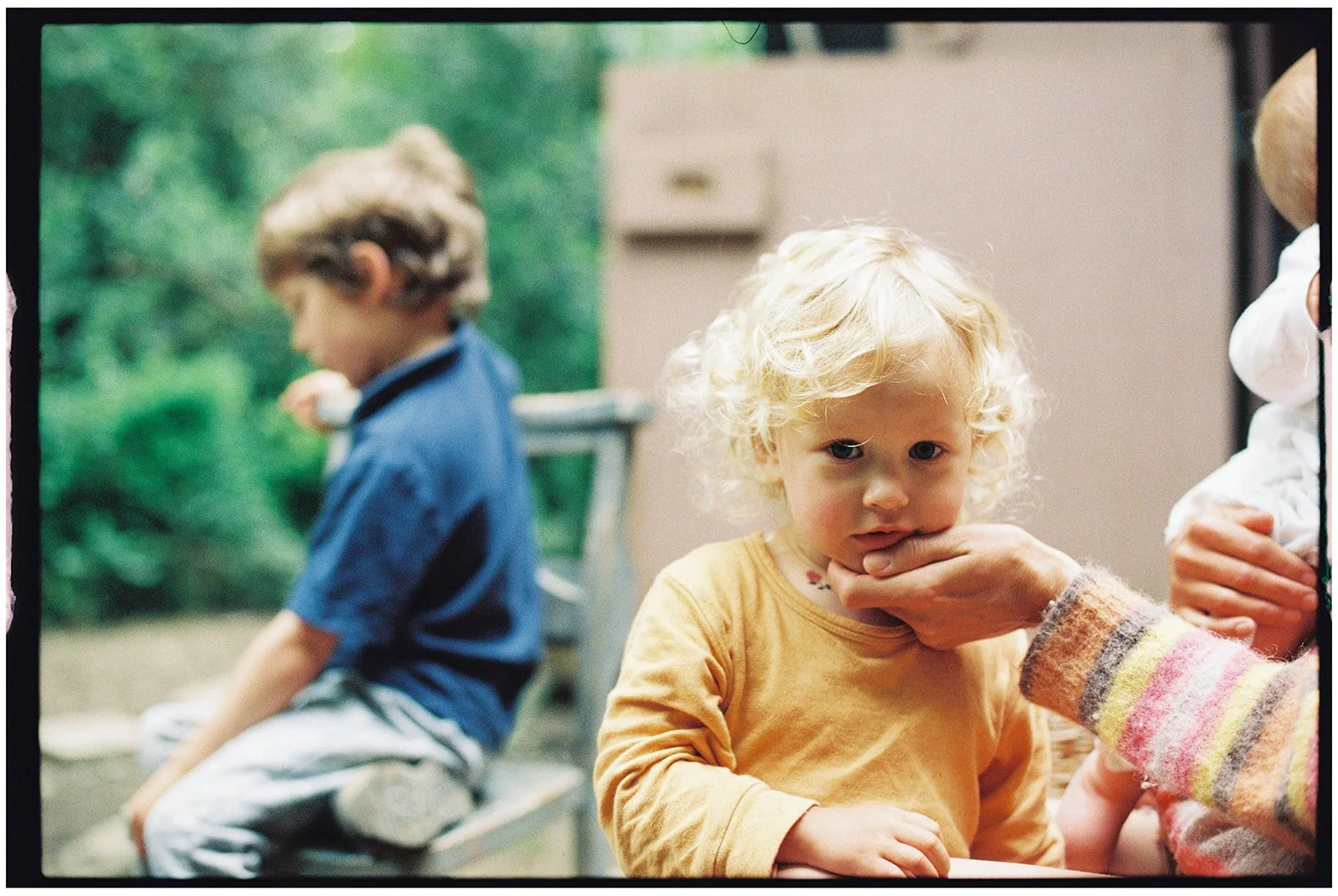 A young blonde child in a yellow shirt looking at the camera, with an adult hand touching their chin. In the background, there is a blurred child sitting on a bench outdoors.