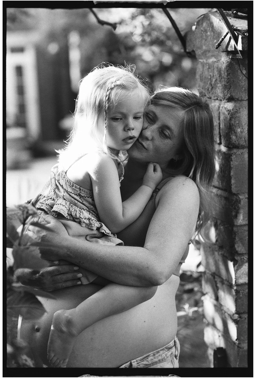 A woman holding a young girl in her arms, standing outdoors near a brick structure, with trees blurred in the background.