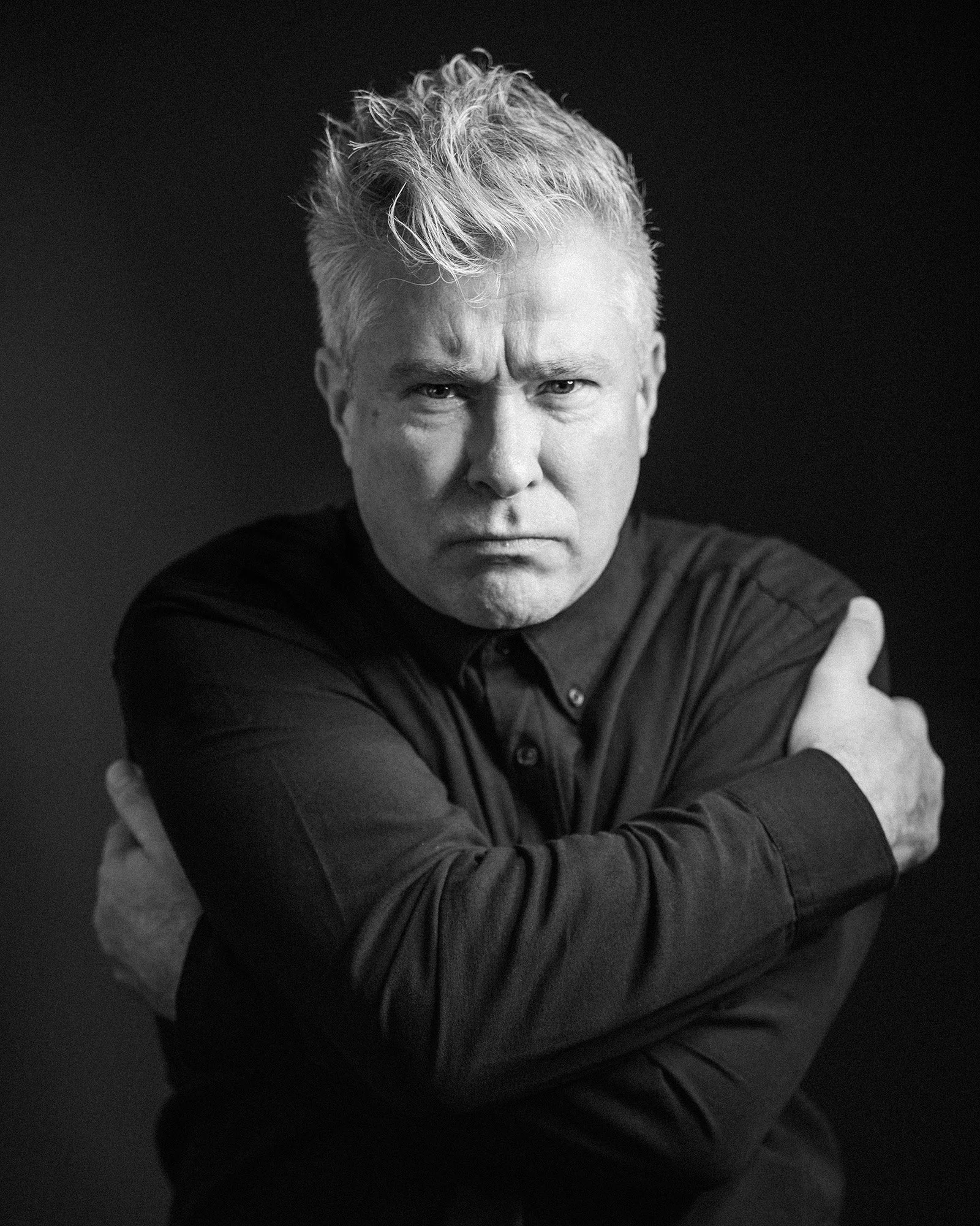 Black and white portrait of a middle-aged man with short, messy hair, wearing a dark shirt, crossing his arms and looking intensely at the camera.