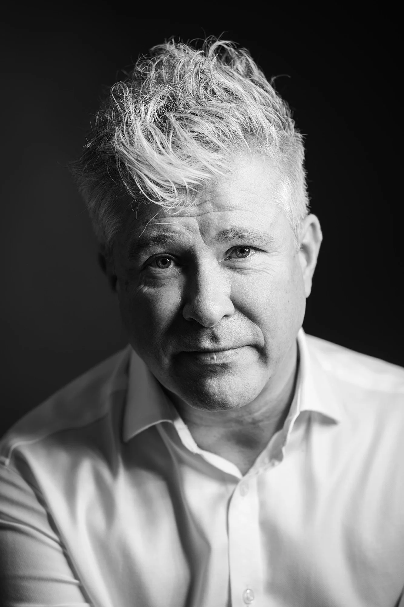 Black and white portrait of a middle-aged man with light-colored, wavy hair, wearing a white shirt, facing the camera with a slight smile.