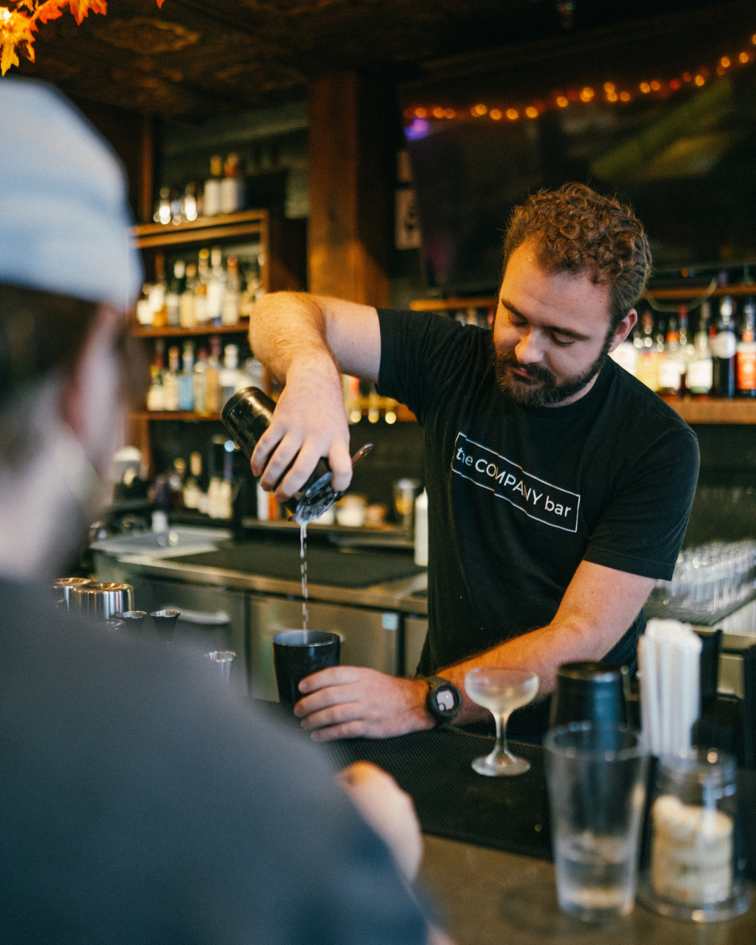 A bartender pouring a drink into a black cup at a bar with bottles of alcohol in the background.