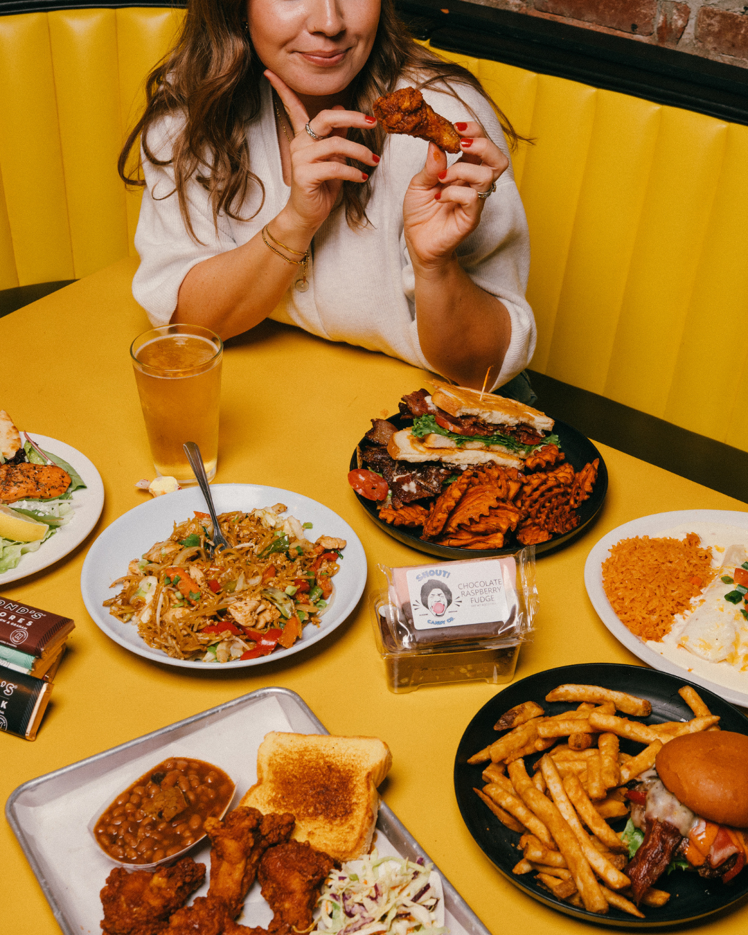 Woman holding a chicken drumstick at a table filled with various dishes including fried chicken, ribs, Thai noodles, rice, fries, and a glass of beer in a restaurant with yellow seating.