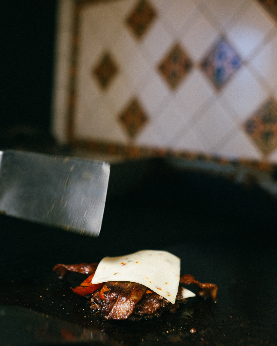 A burger patty being grilled with toppings including a slice of cheese.
