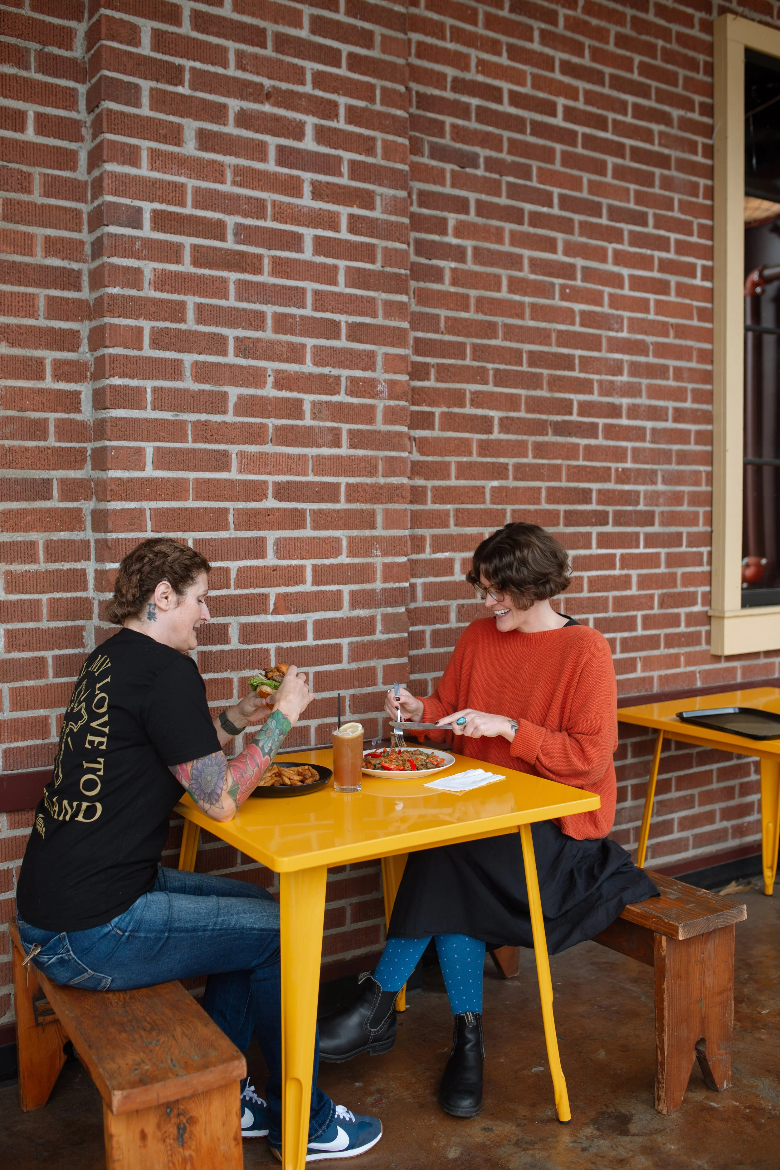 Two people sitting on an outdoor patio on benches at a yellow table with a brick wall behind them enjoying a burger, fries, and a Thai noodle dish