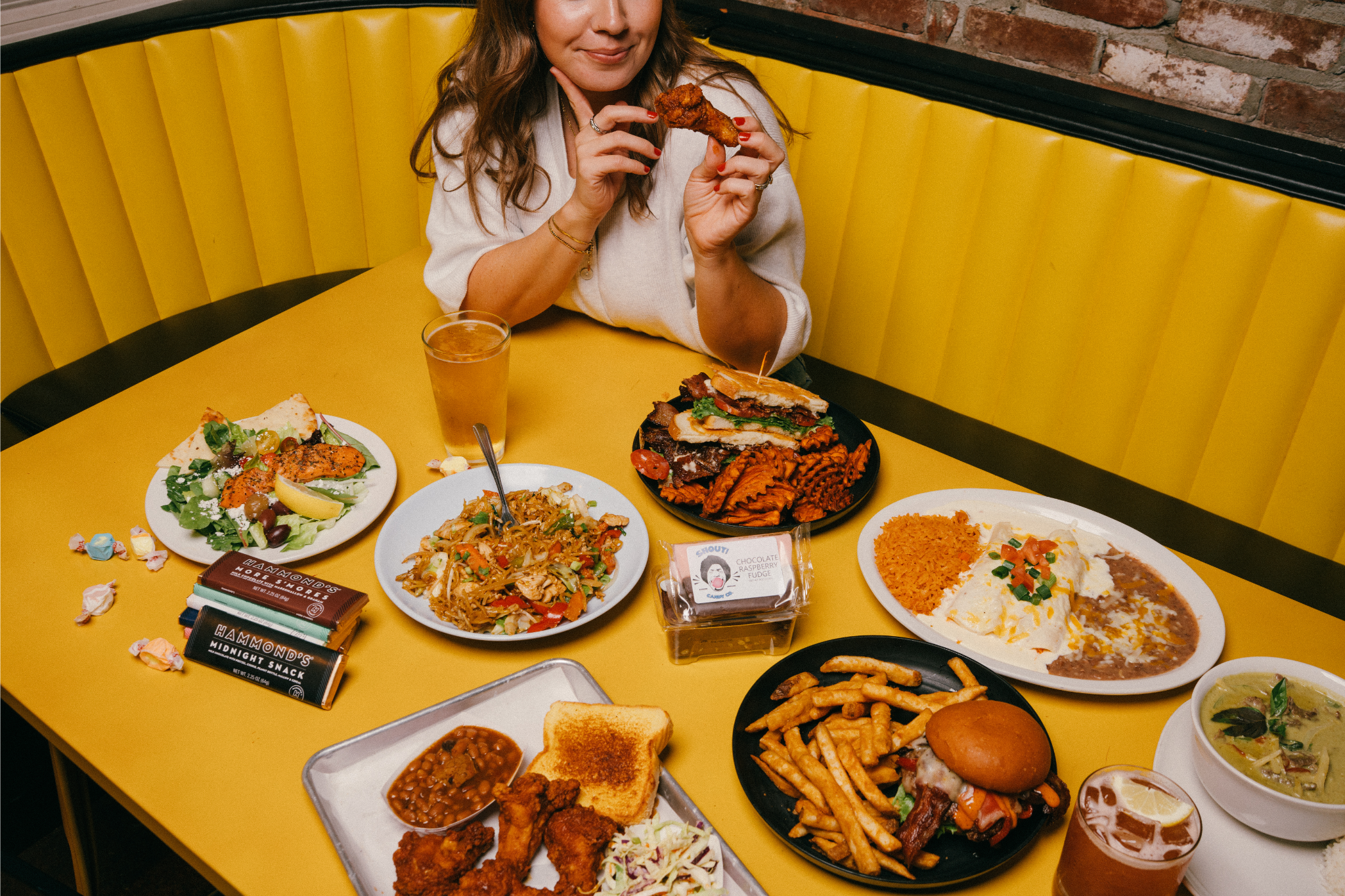 A woman sitting at a yellow booth enjoying many dishes including fried chicken, Thai noodles, nachos, a burger, and drinks in a restaurant.