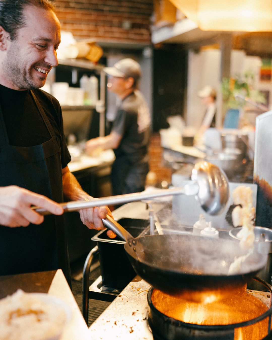 A chef smiling while cooking in a kitchen, using a spatula to fry food in a black skillet over a stove with a red-orange flame.