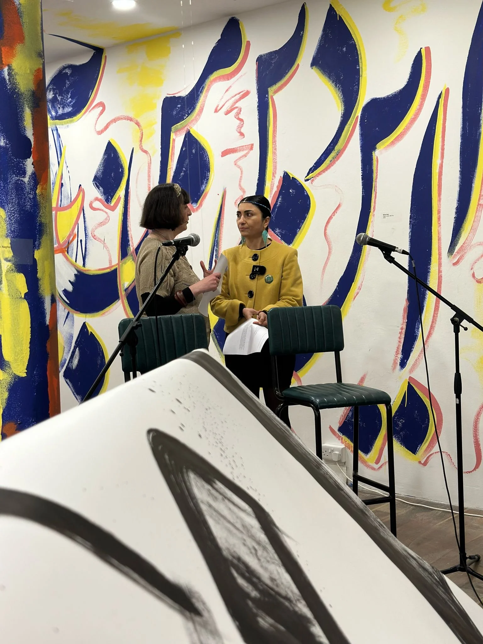 View from behind a large, hanging, suspended black and white scroll to where Sahar and Mab are chatting onstage at the opening event.jpeg