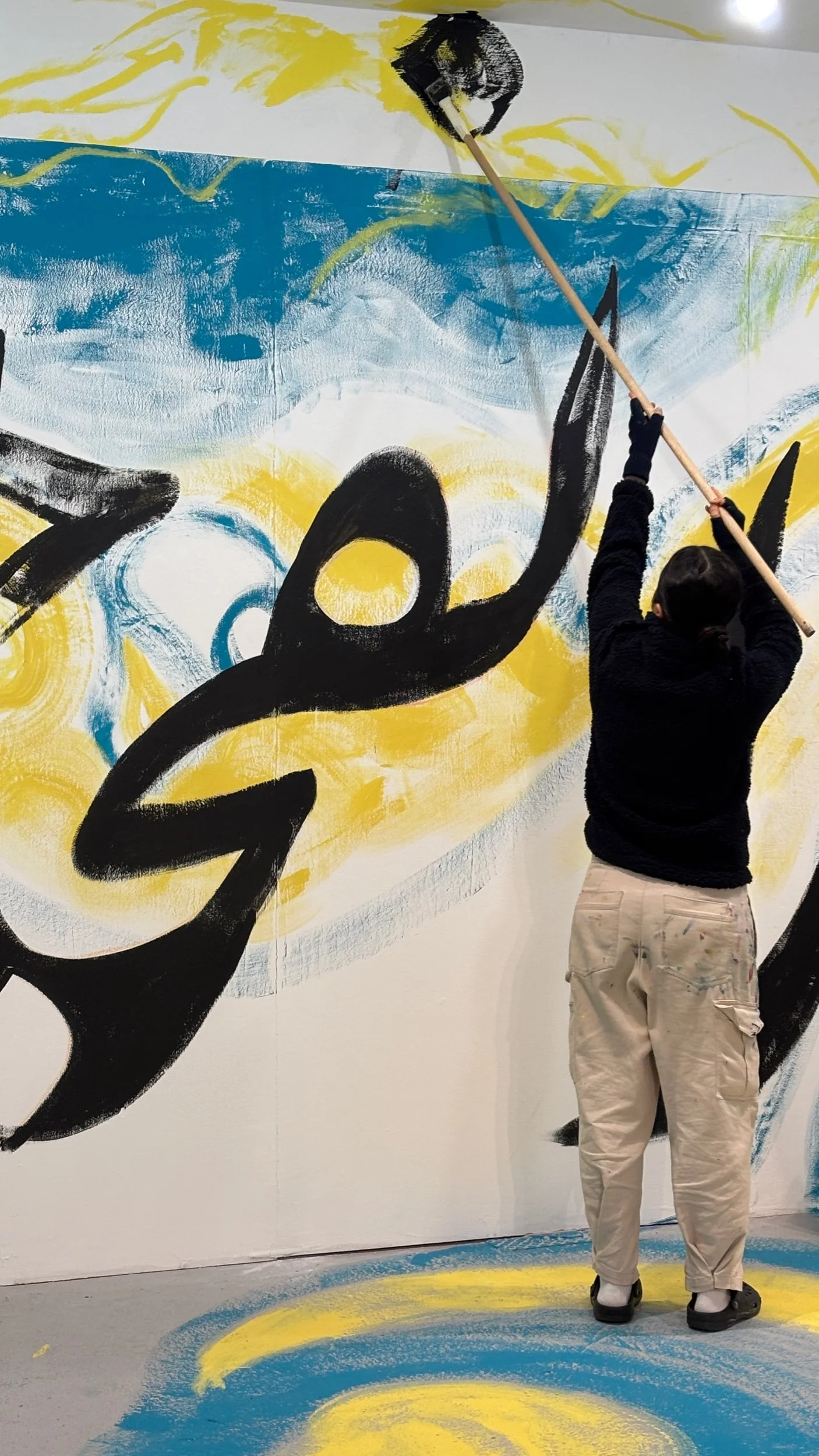 Sahar from behind, standing and stretching with a long brush to paint a gallery ceiling and wall.jpeg