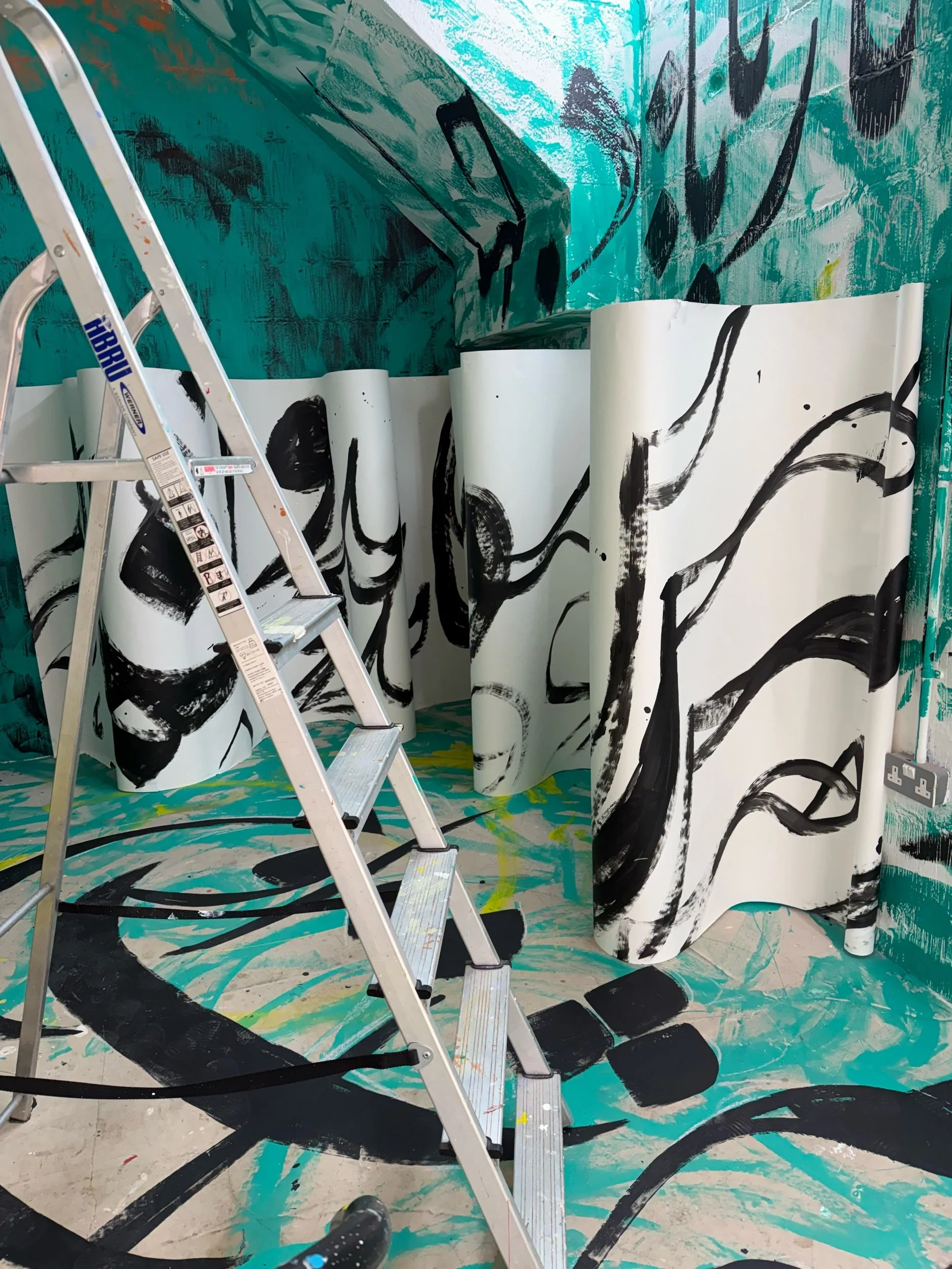 A large white scroll with black calligraphy across it sits in the gallery space with black calligraphy on the turquoise walls behind.jpeg