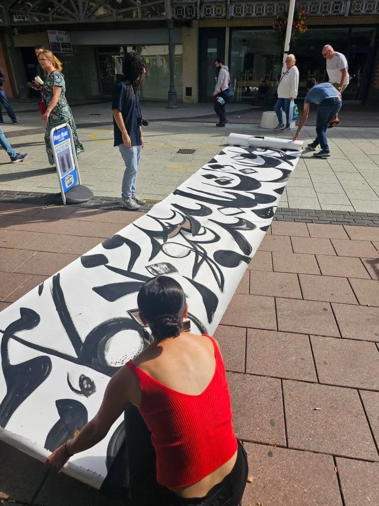 Sahar squats outside in sunshine, back to camera, as she paints a long, unrolled black and white scroll on the pavement.jpeg